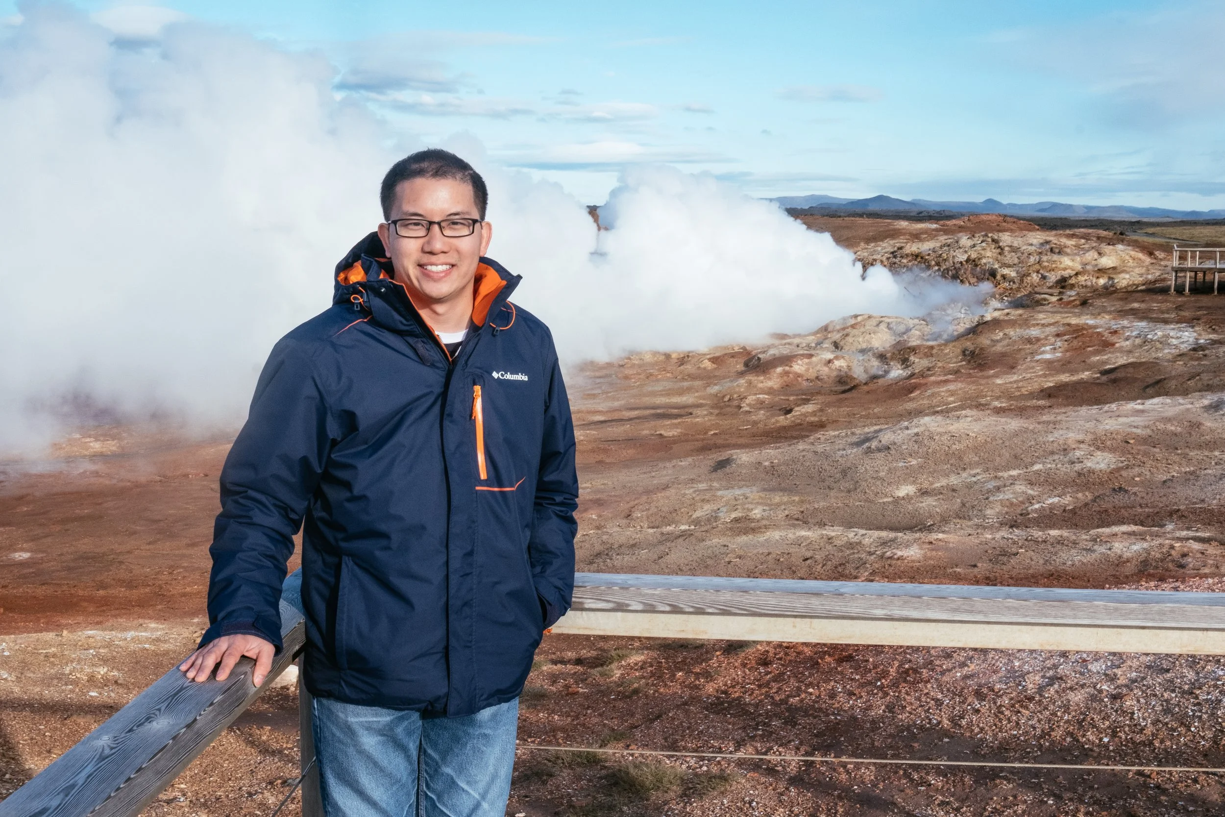A smiling man wearing glasses and a navy jacket standing outdoors near steam and geothermal terrain with mountains in the background.