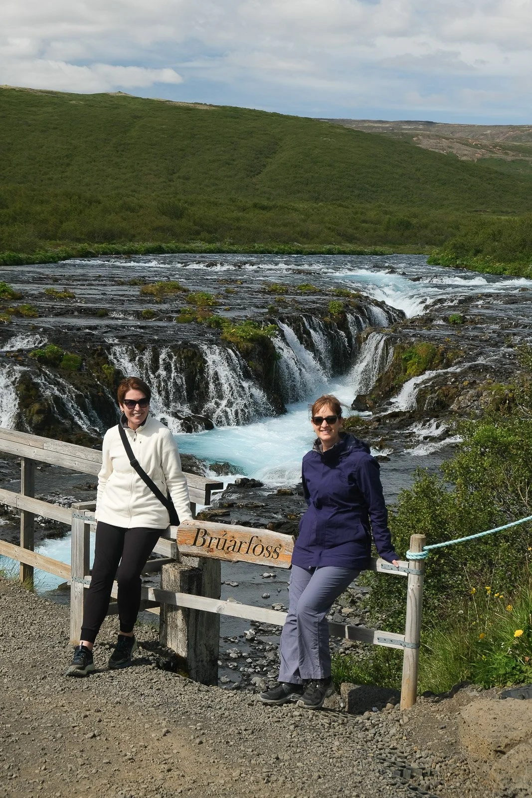 Two women standing on a wooden path at Bruarfoss waterfall with green hills and a cloudy sky in the background.