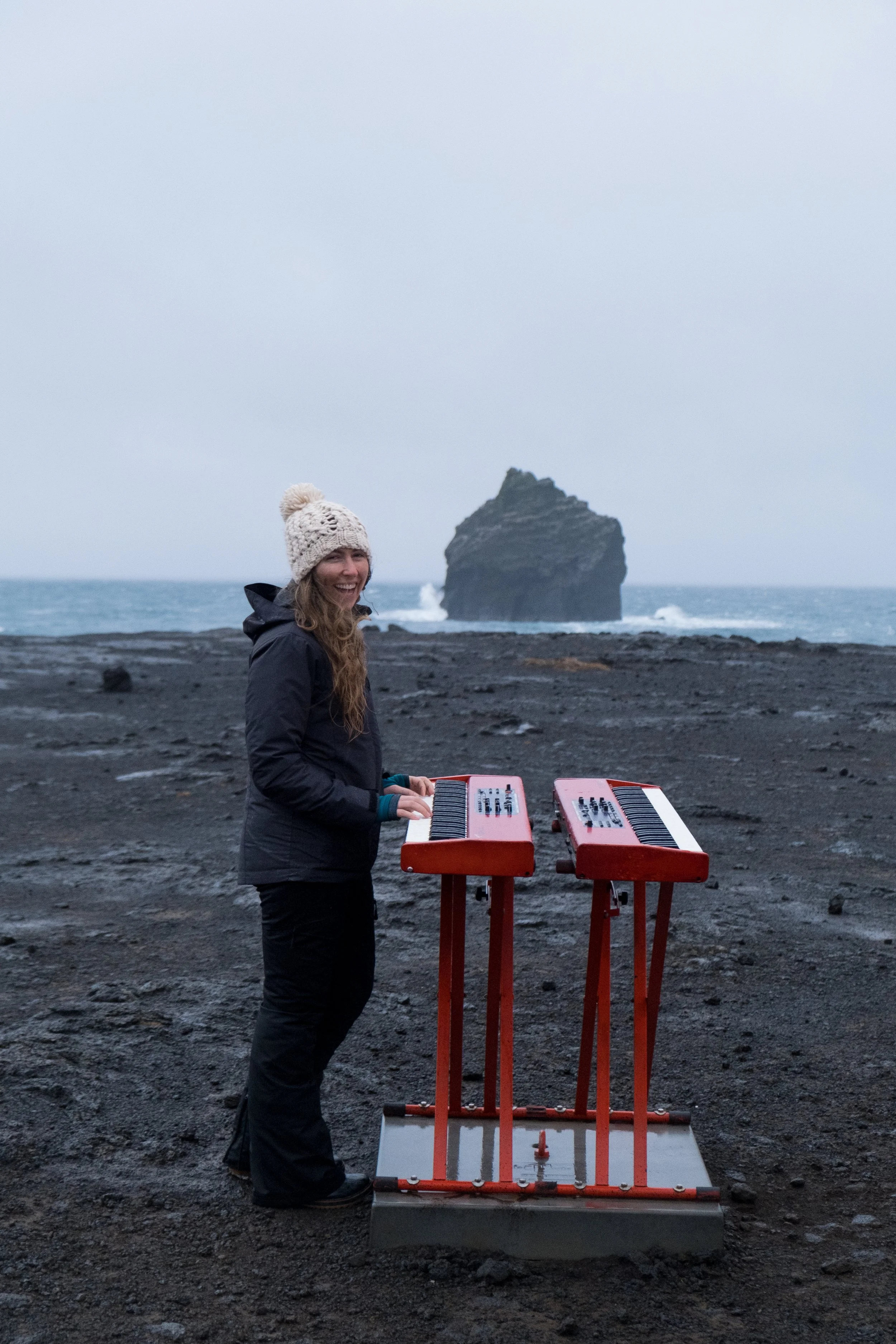 A woman standing outdoors on dark volcanic soil at the beach, wearing a knitted hat and jacket, with a portable electronic keyboard on a stand in front of her. The ocean and a large rock formation are visible in the background.