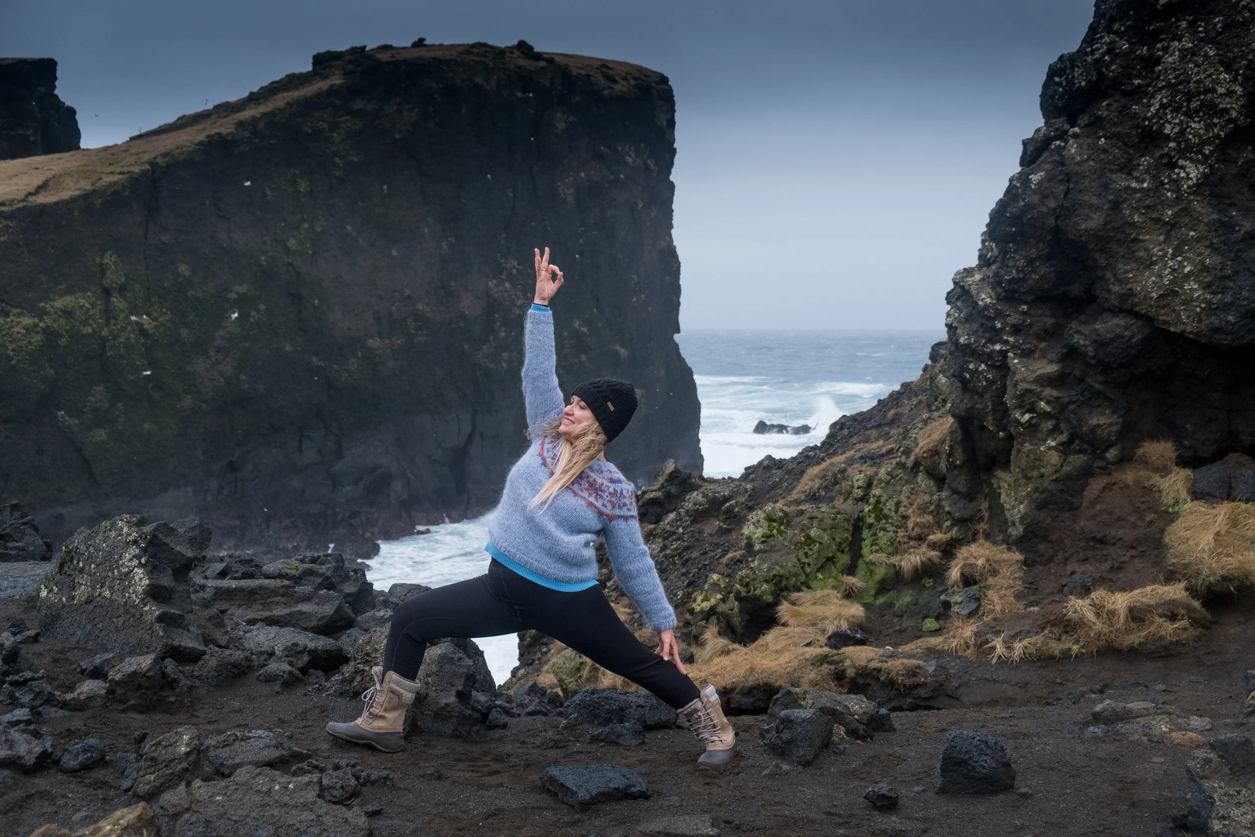 A woman performing a yoga pose on rocky terrain near the ocean, surrounded by cliffs and overcast sky.