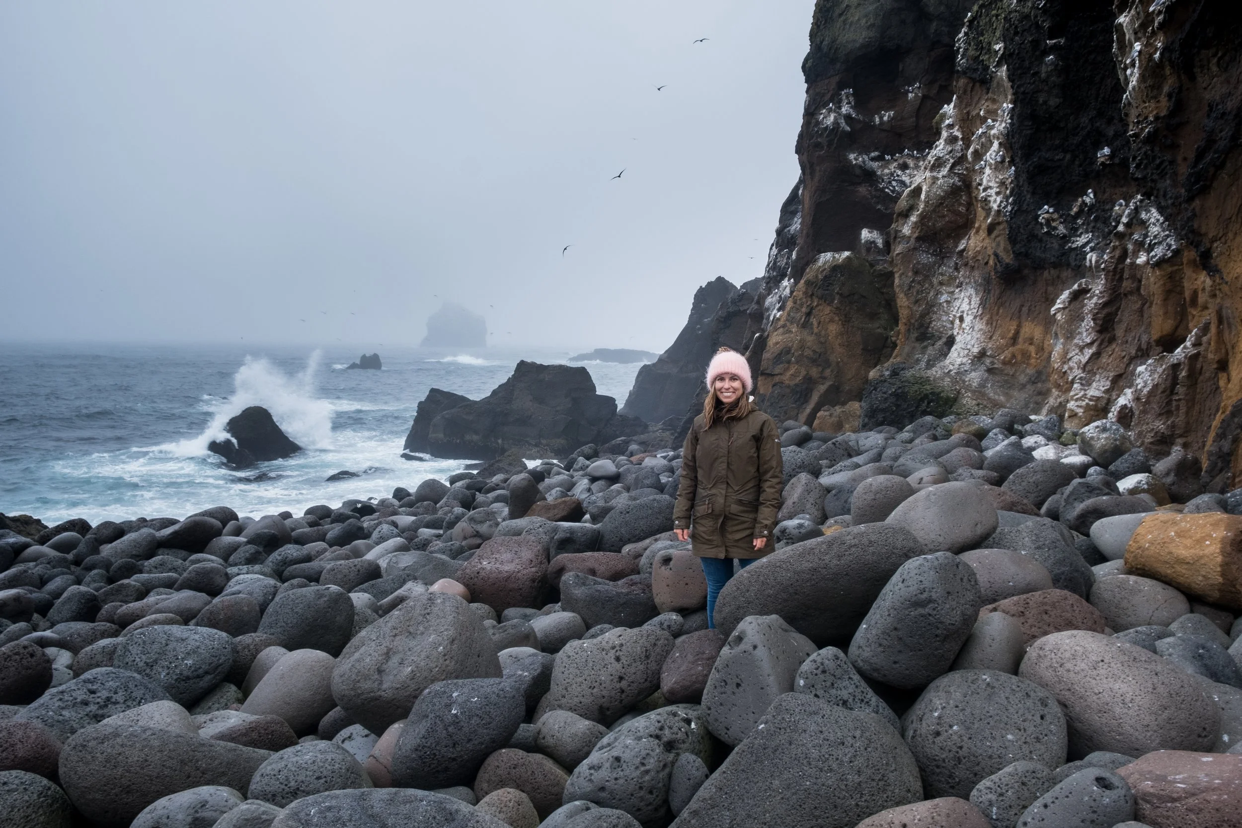 A woman in a brown jacket and pink knit hat standing on a rocky beach with large stones, with cliffs and the ocean in the background, under a cloudy sky.