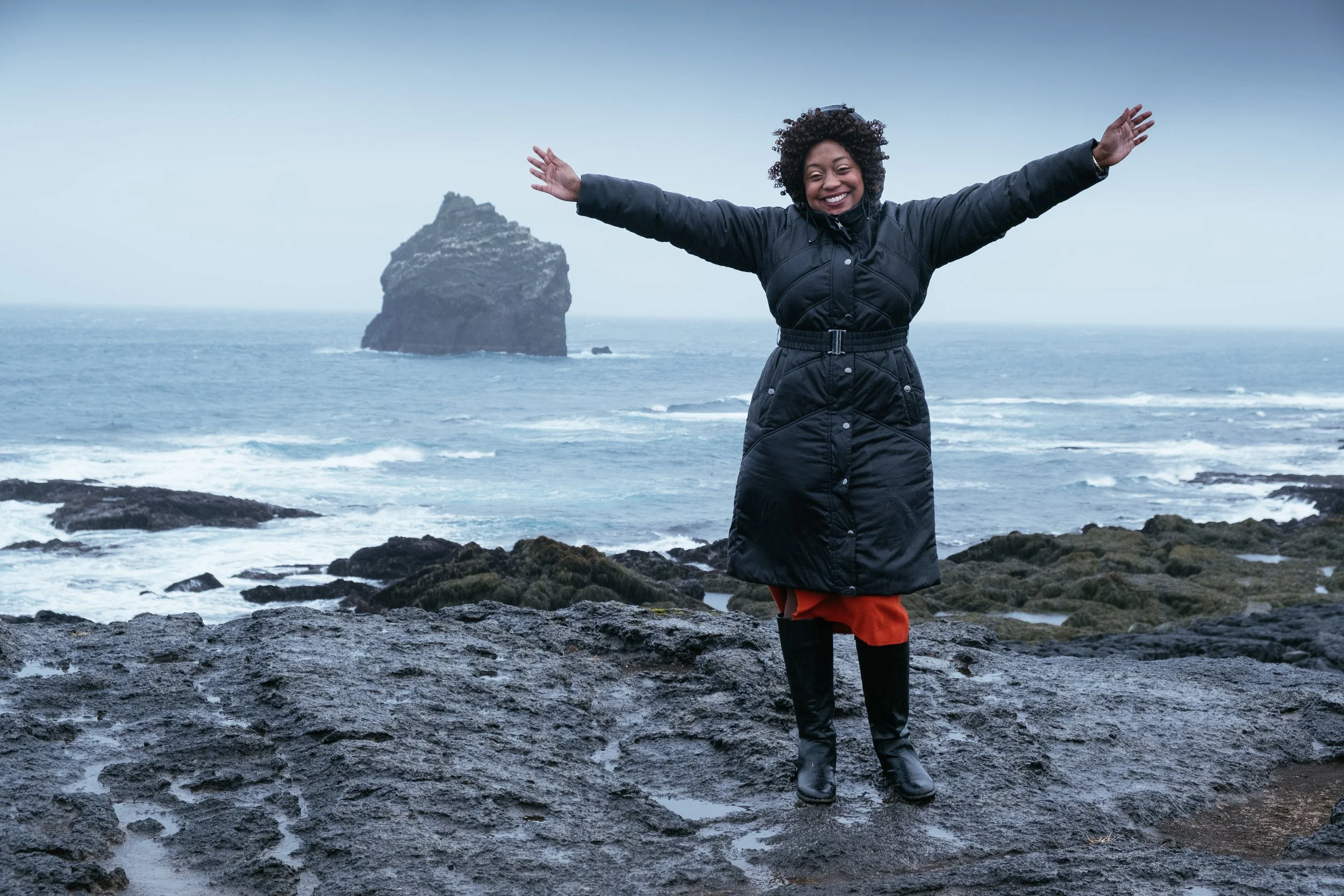 A woman standing on rocky shoreline with arms outstretched, smiling, wearing a black jacket, red pants, and black boots, with ocean and large sea stack in the background.