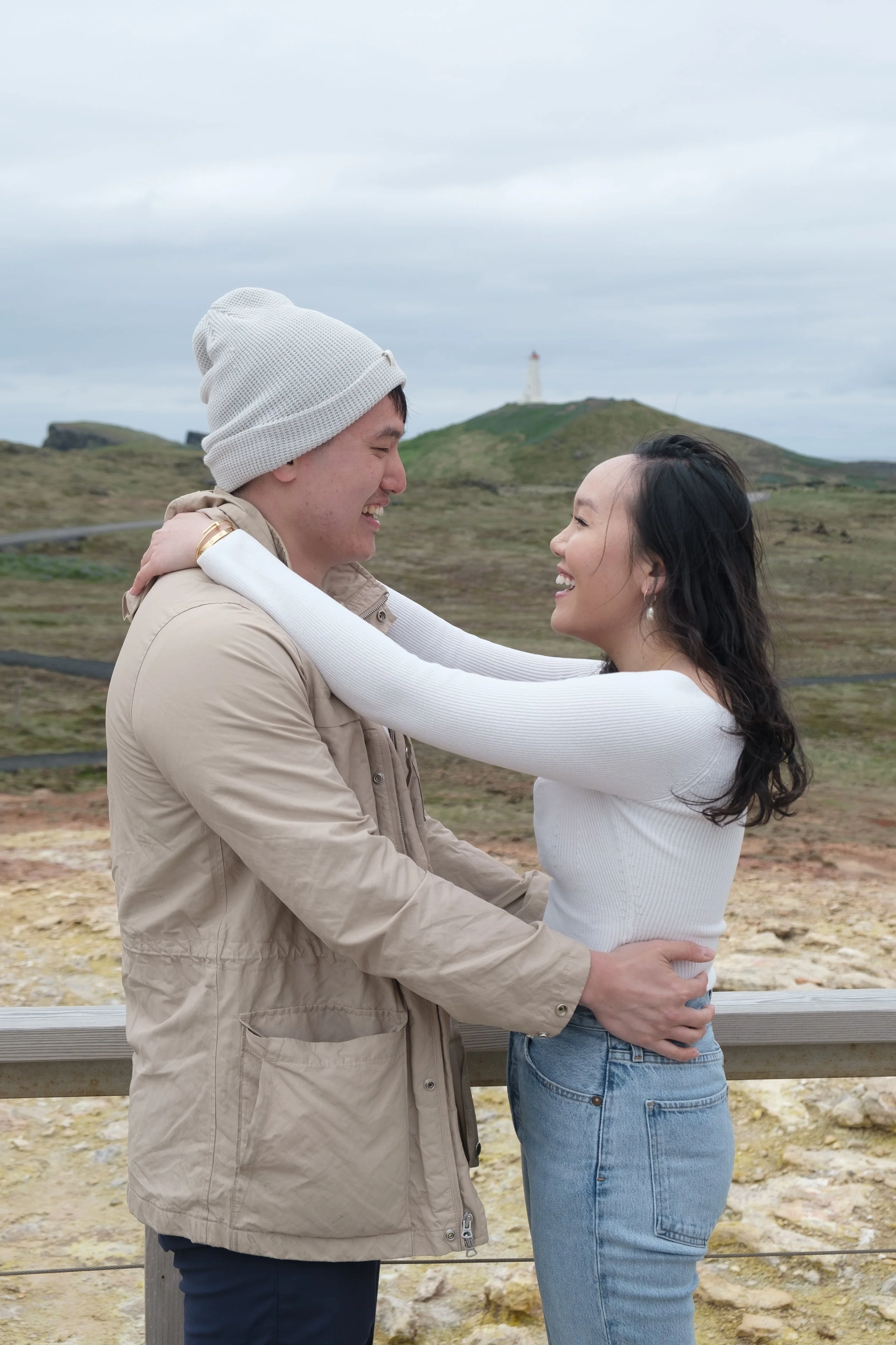 A man and woman are embracing outdoors, smiling and looking at each other, with a landscape of grass, hills, and a lighthouse in the background.