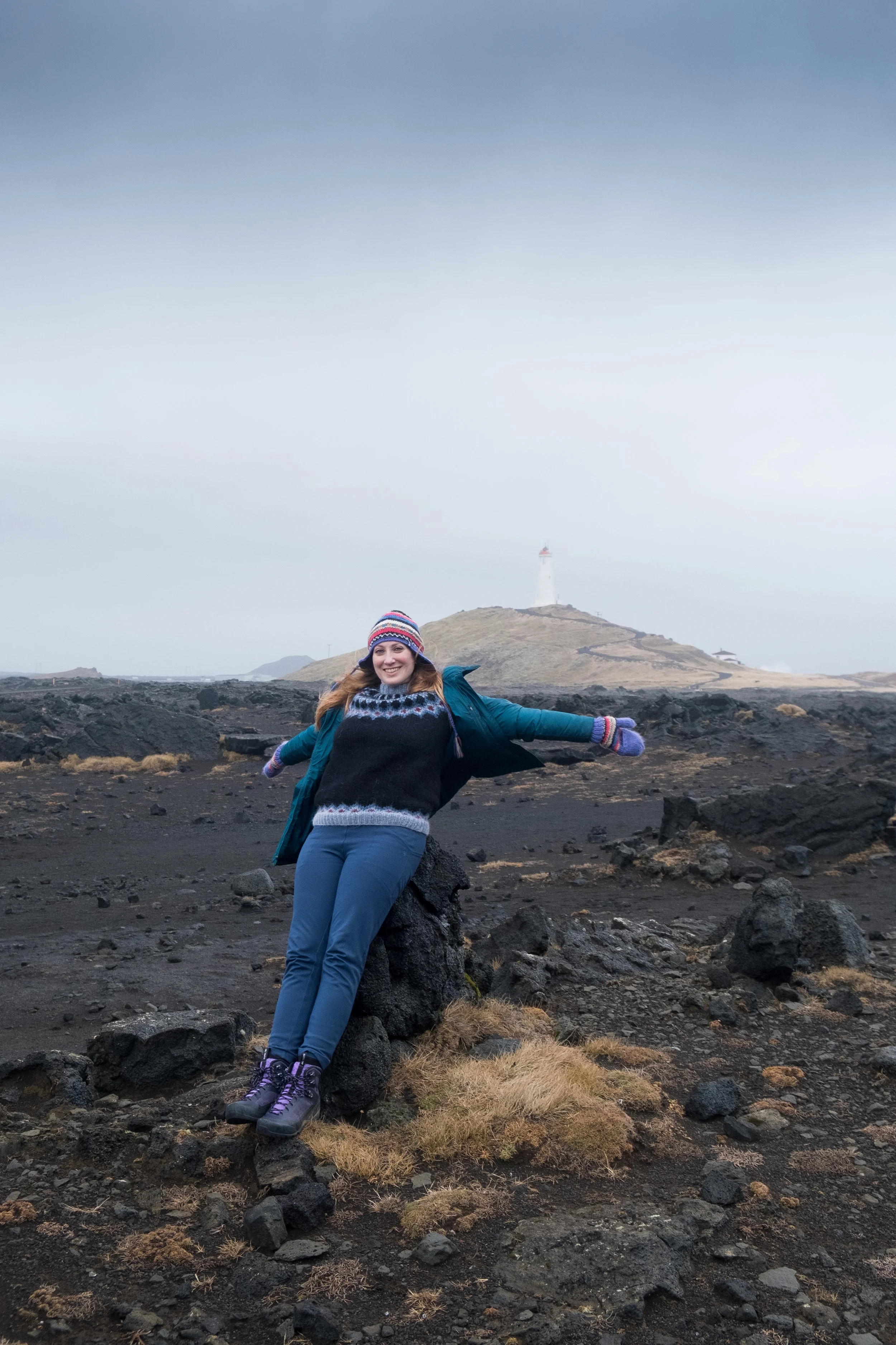 A woman smiling and posing with arms outstretched on rocky volcanic terrain, wearing a colorful winter hat, coat, and boots, with a lighthouse on a hill in the background under overcast sky.