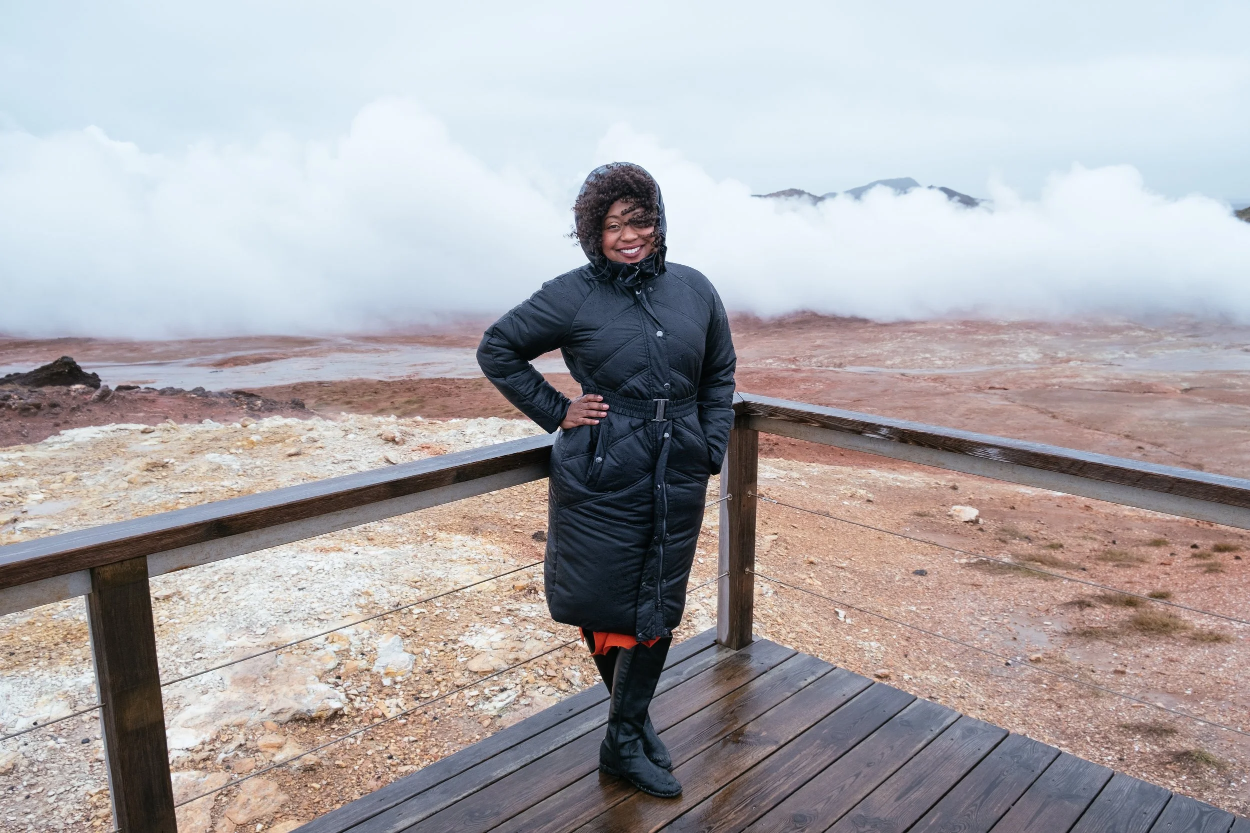 A woman standing outdoors on a wooden platform, wearing a black jacket and smiling. The background features a cloudy sky and volcanic landscape with steam or mist rising from the ground.