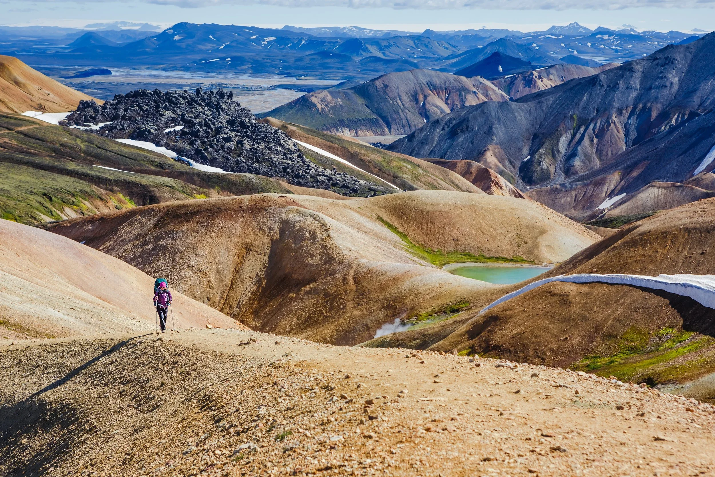 A hiker with a walking stick walking on a dirt trail through colorful volcanic mountains with patches of snow, a hot spring, and distant peaks under a partly cloudy sky.