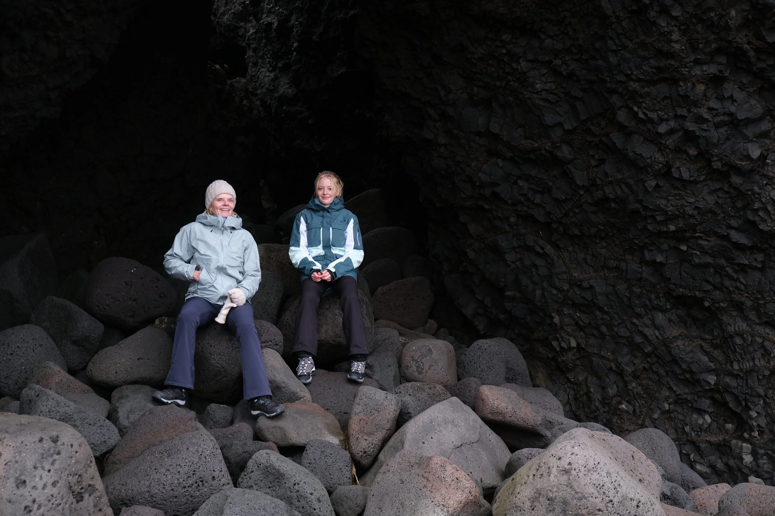 Two women sitting on large rocks inside a dark volcanic cave with rocky, textured walls.