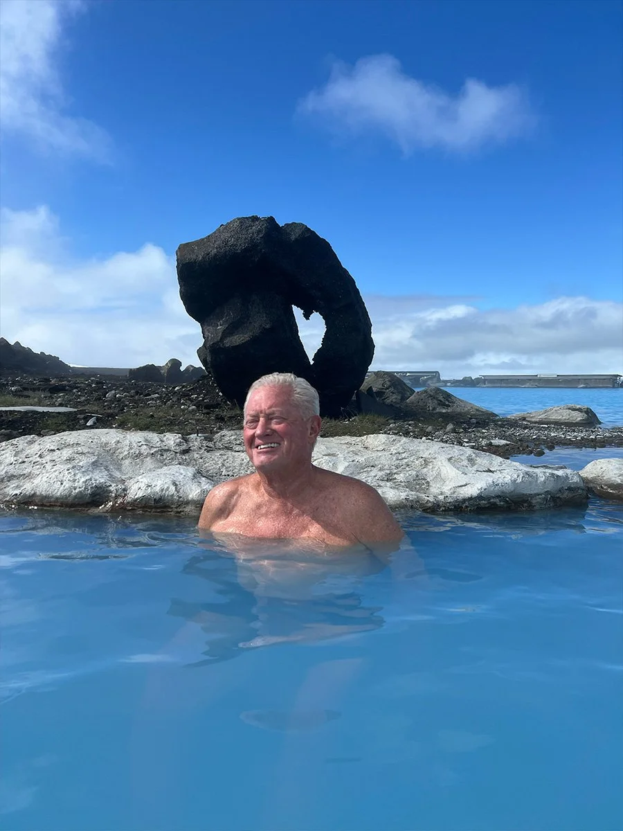 An elderly man swimming in a natural hot spring or pool outdoors with a large, abstract rock formation and a partly cloudy blue sky in the background.