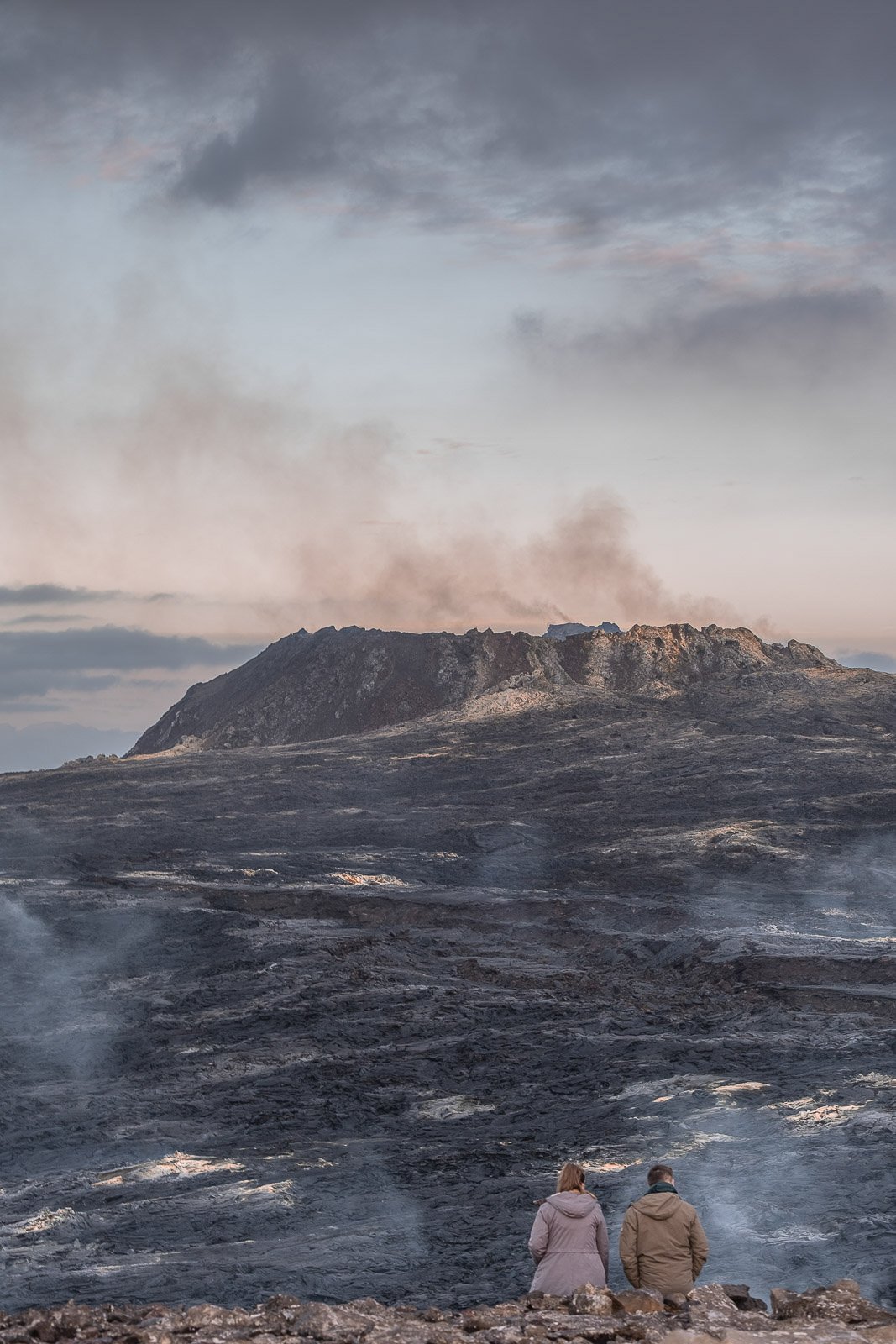 Two people sit on rocks facing a volcanic landscape with smoke rising from the volcano, dark rocky terrain, and a cloudy sky in the background.