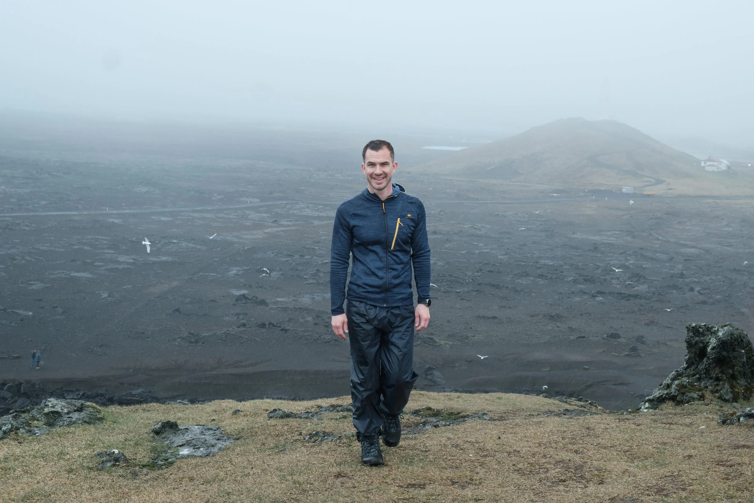A man standing outdoors in a misty, mountainous landscape with volcanic soil, wearing a blue jacket and black pants.