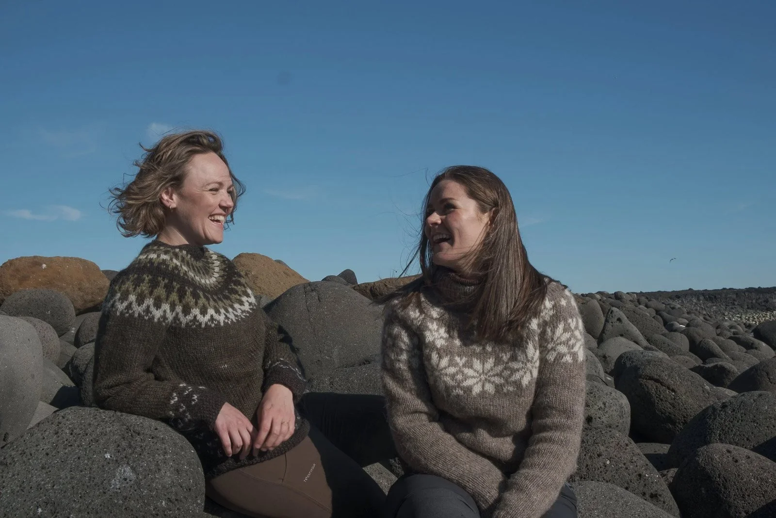 Two women wearing patterned sweaters sitting on rocks outdoors, smiling and laughing at each other.