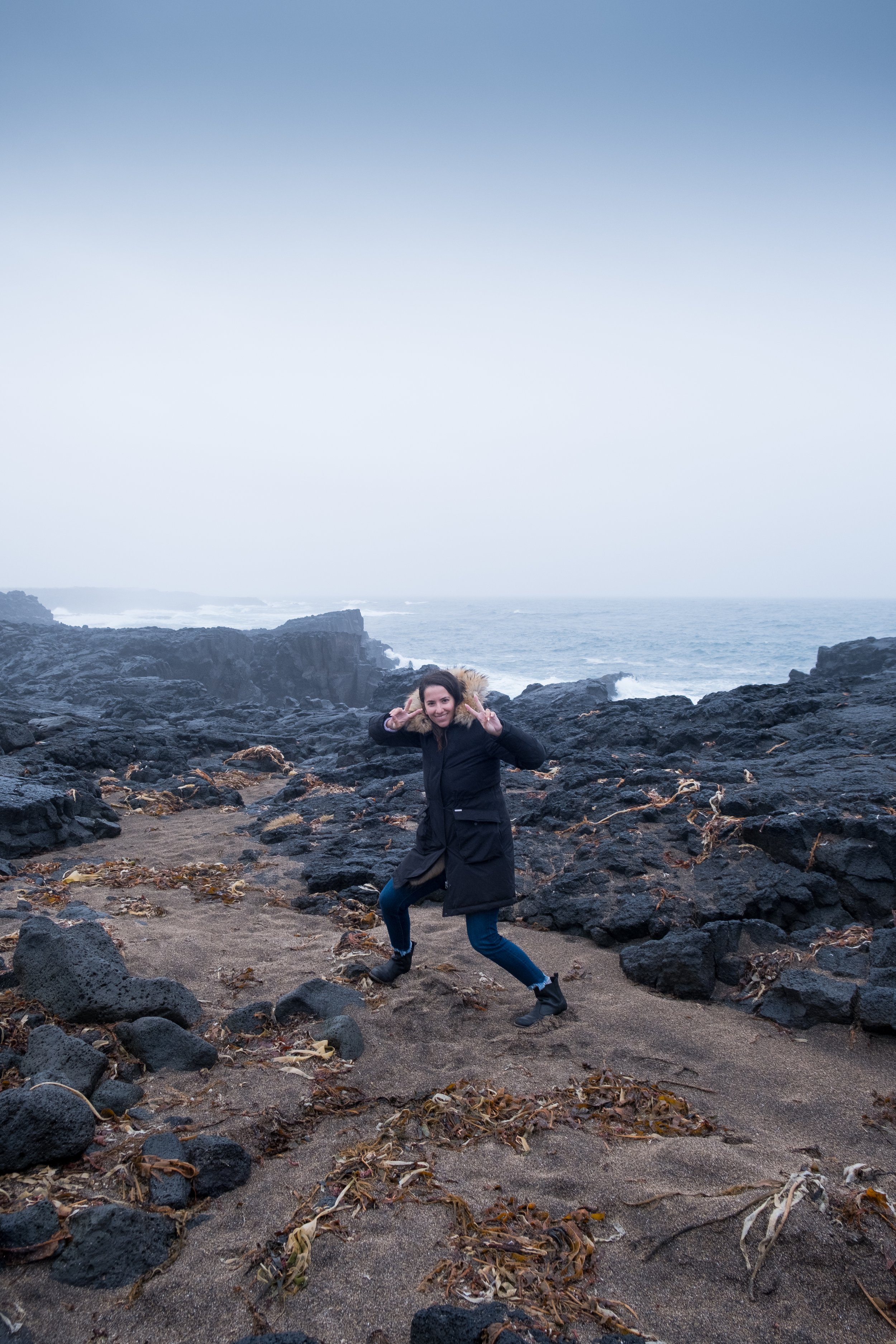 A woman in a black winter coat and jeans making a peace sign with both hands while standing on a rocky and sandy beach with dark rocks, dried seaweed, and an ocean in the background under a cloudy sky.