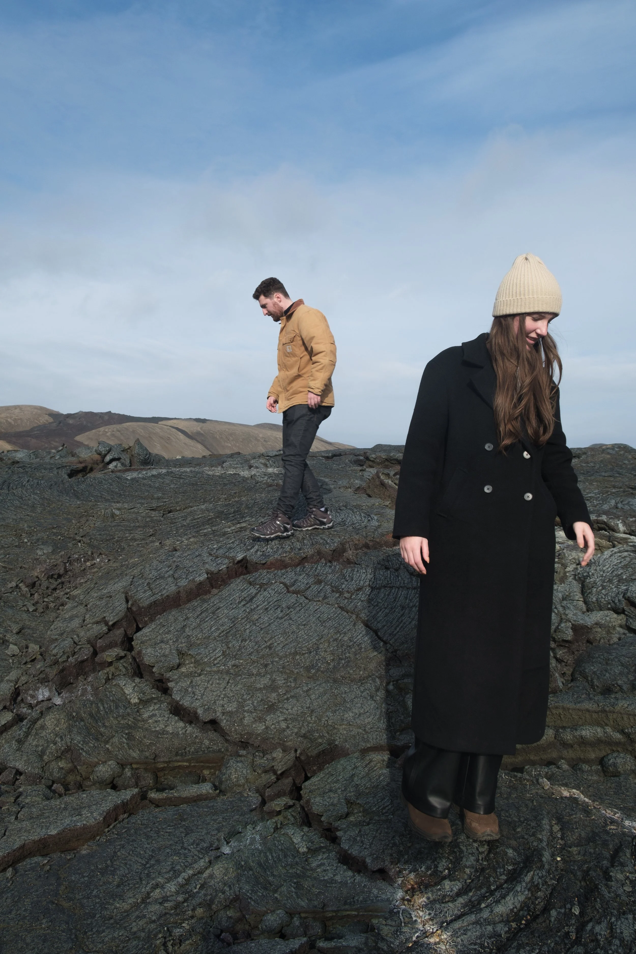 Two people standing on a rocky landscape under a partly cloudy sky. One person is a woman in a black coat and a beige knit hat, looking down. The other person is a man in a tan jacket, looking down at the ground.