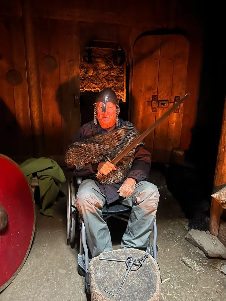 A person dressed as a knight with chainmail and a helmet, sitting in a wheelchair and holding a sword, inside a wooden room with a fierce expression.