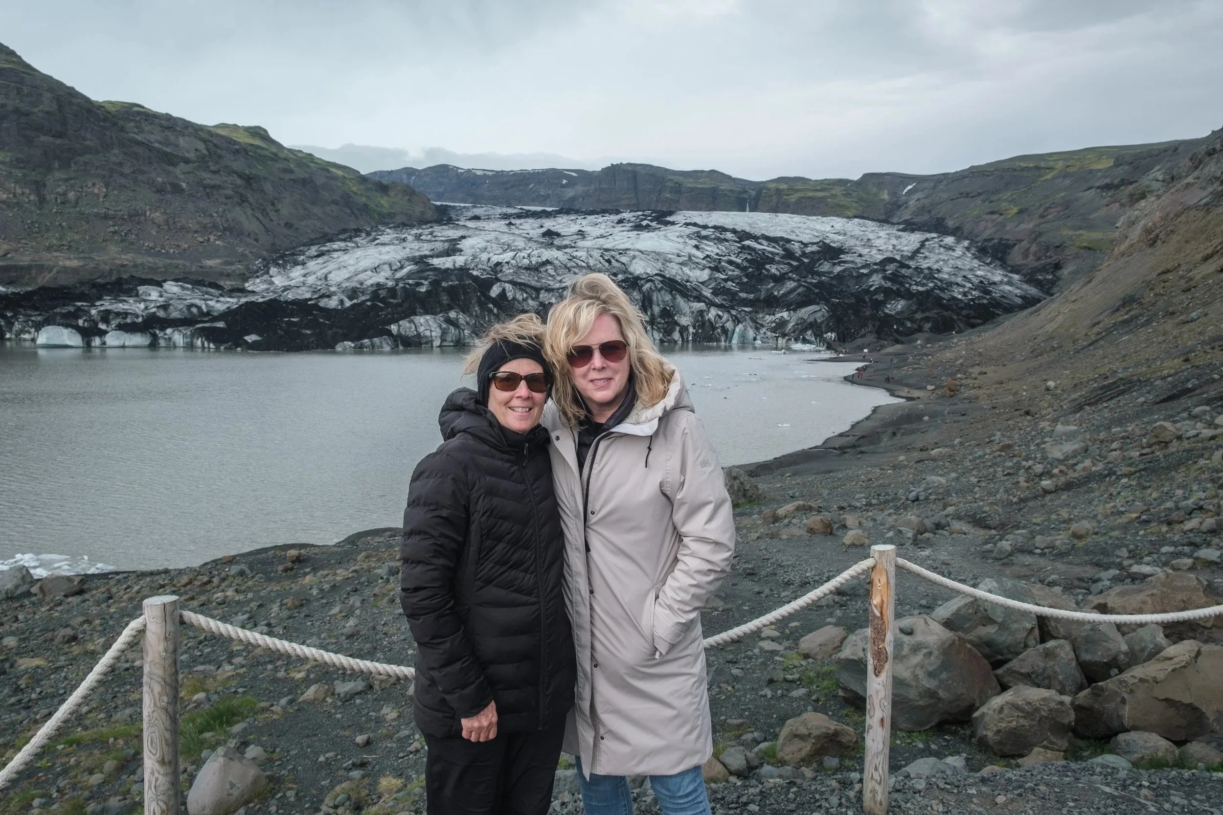 Two women standing outdoors near a body of water with a glacier and mountainous terrain in the background, wearing jackets and sunglasses.