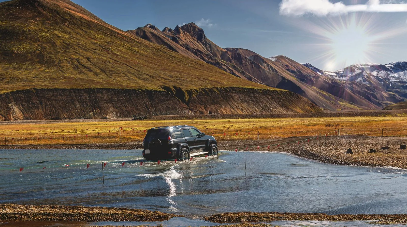 A black SUV driving through a shallow body of water in a valley with mountains in the background, under a bright sun in a clear sky.