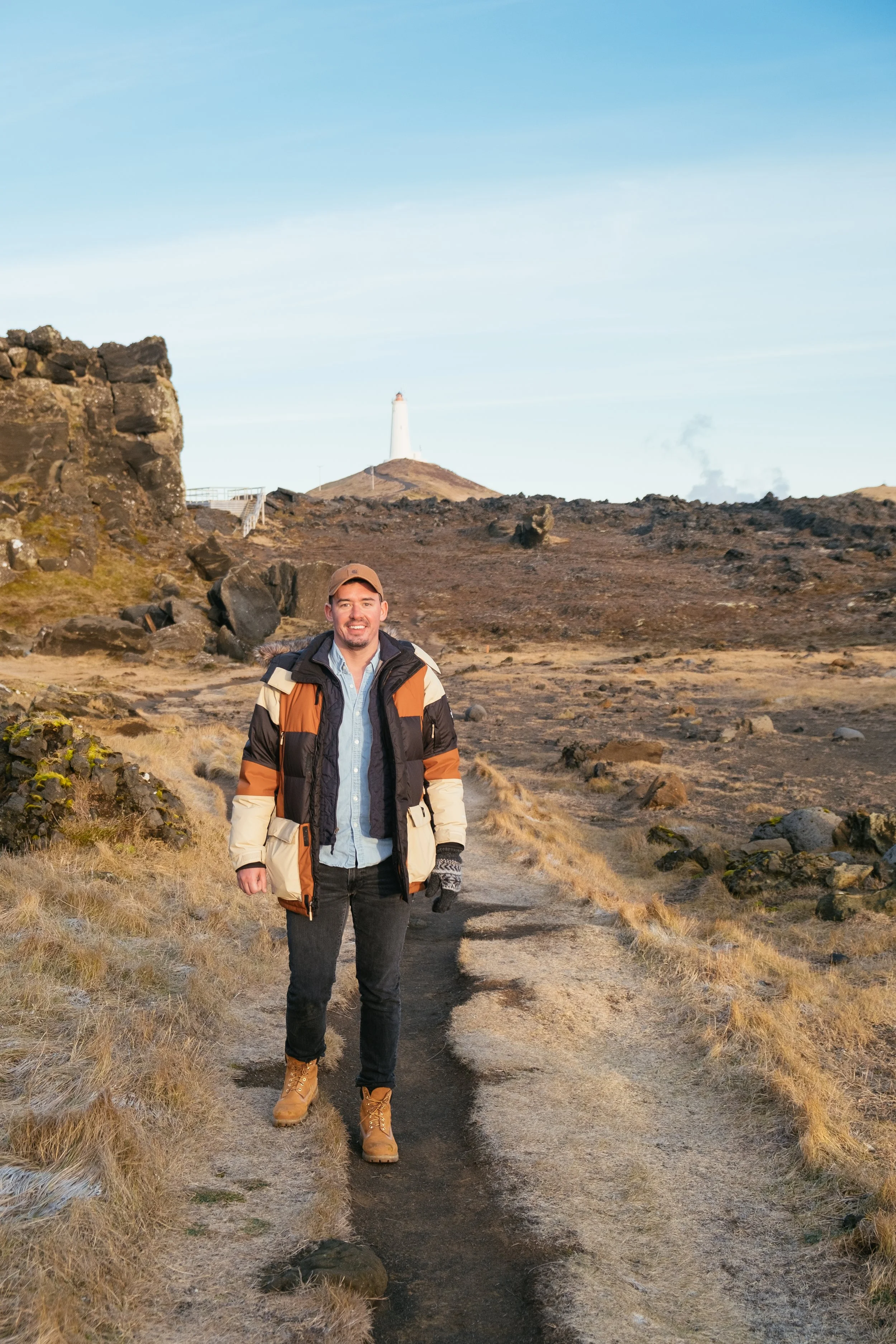 A man wearing a beige, black, and orange jacket, jeans, tan boots, and a brown cap walking on a dirt trail in a volcanic landscape with brown rocks and yellow grass, with a lighthouse on a hill in the background under a clear sky.