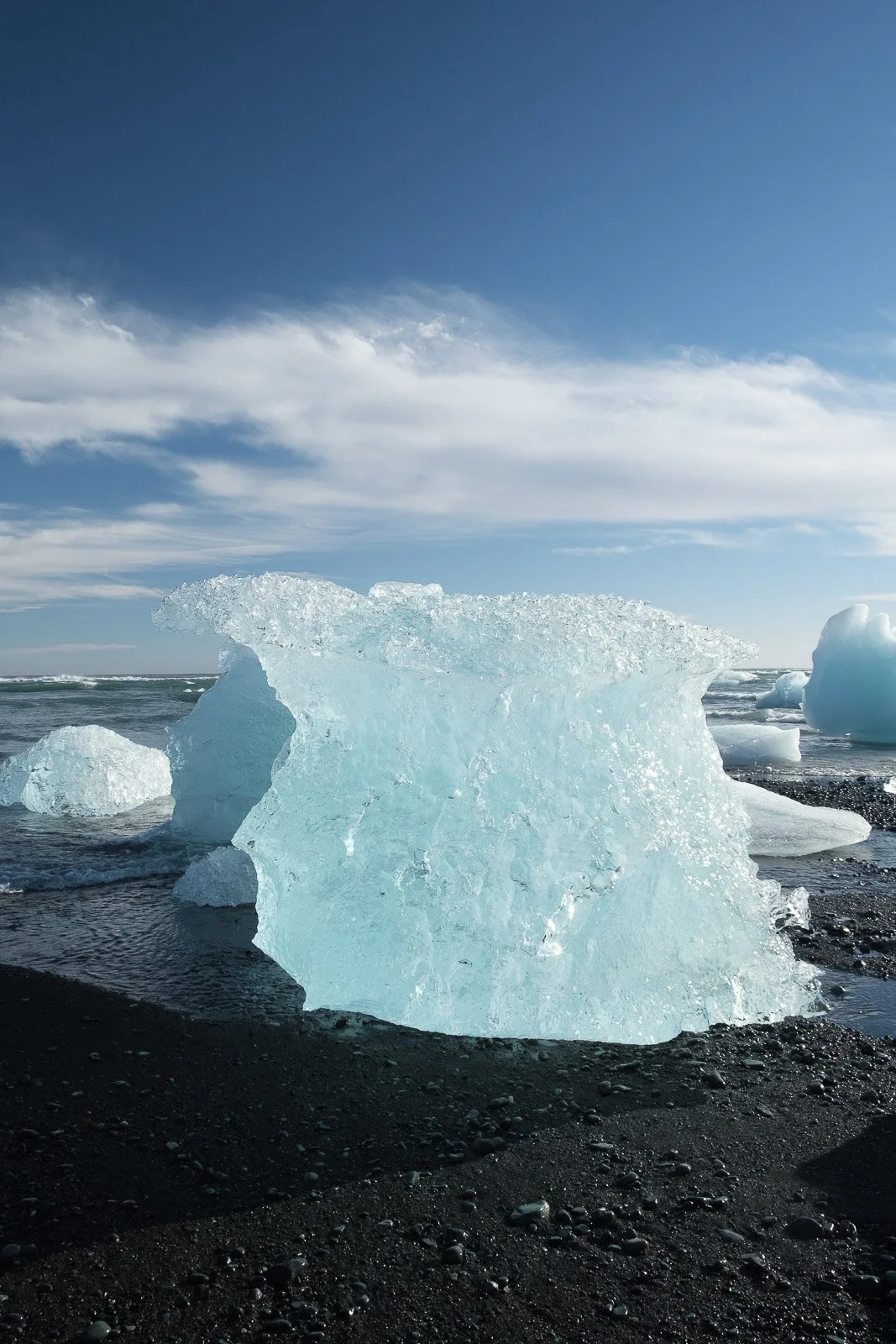 Large floating icebergs on a black sandy beach under a partly cloudy sky.