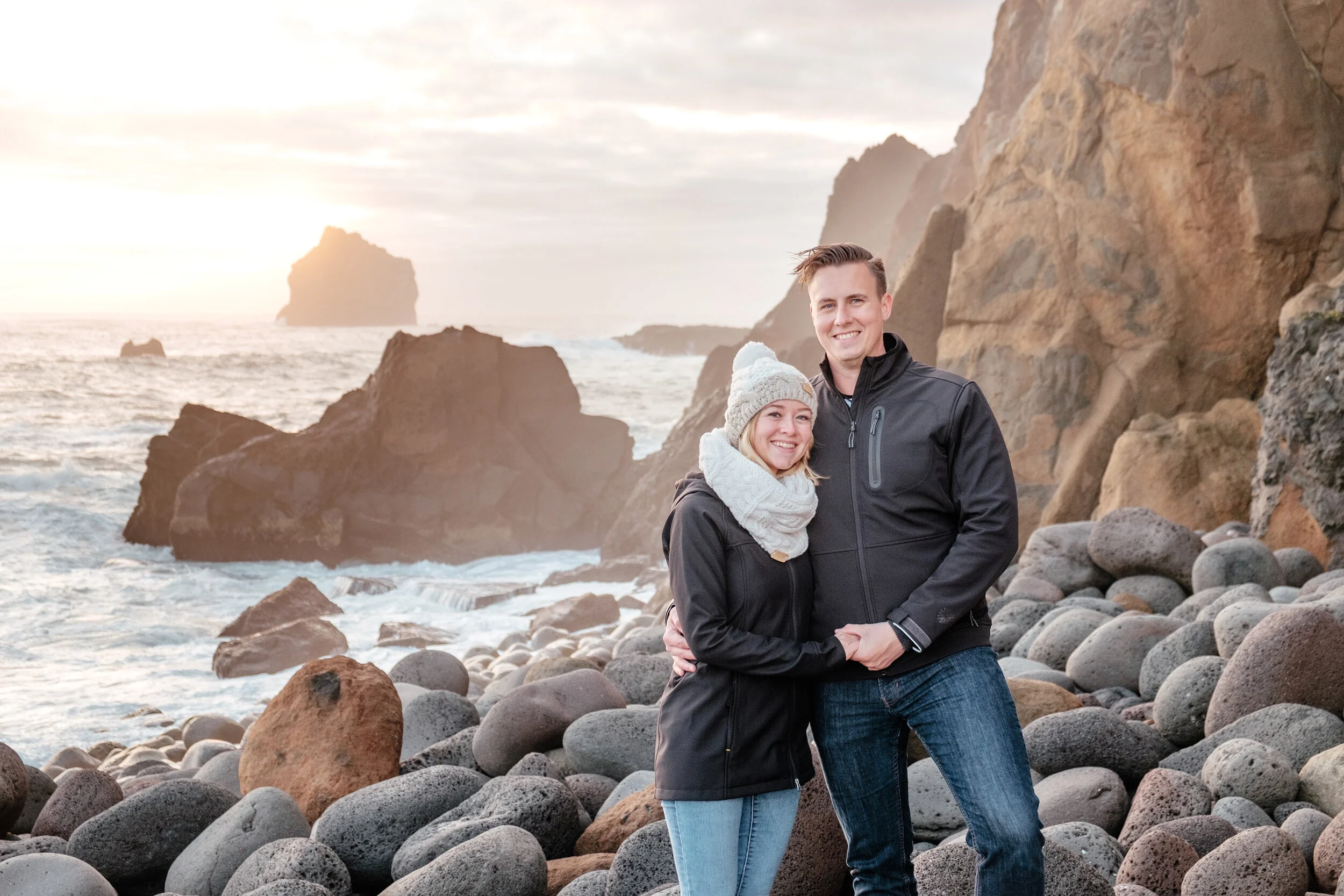 A smiling couple standing on a rocky beach with large rocks and cliffs in the background, during sunset.