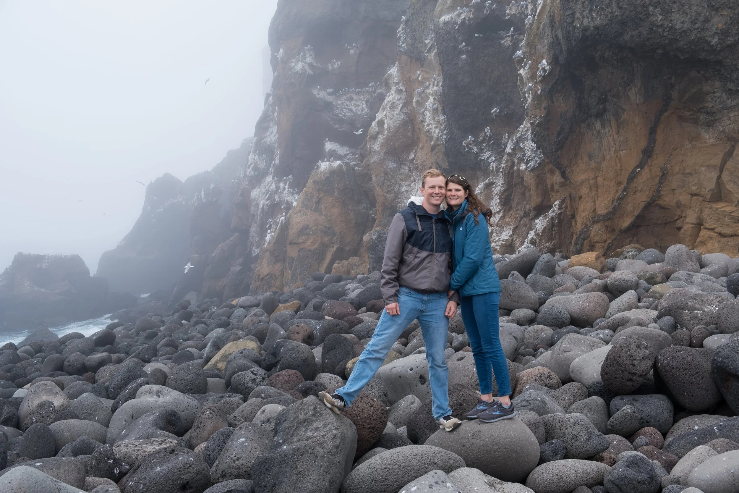A couple stands together on a rocky beach with large stones, smiling at the camera. Behind them are high cliff faces and a misty, foggy atmosphere, creating a scenic and rugged coastal landscape.
