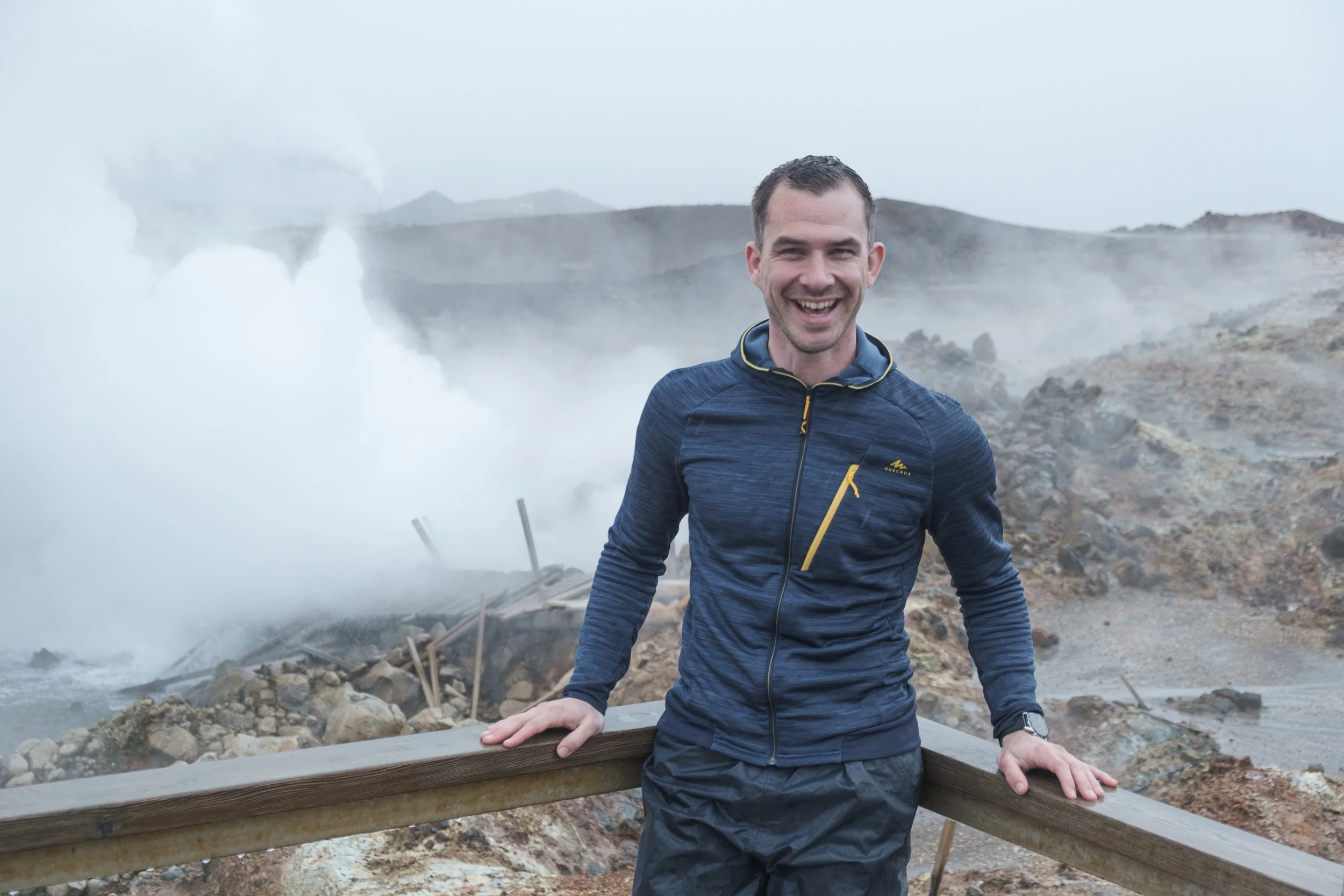 A smiling man in a blue jacket leaning on a wooden railing at a geothermal area with steam rising from the ground.