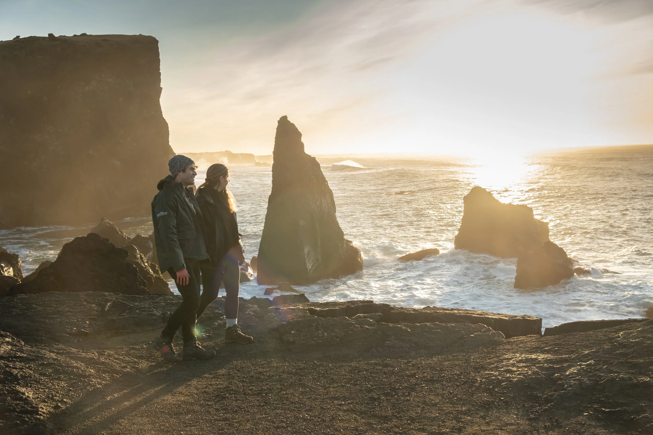 Two people standing on rocks on a beach near large sea stacks during sunset, wearing jackets and hiking shoes.