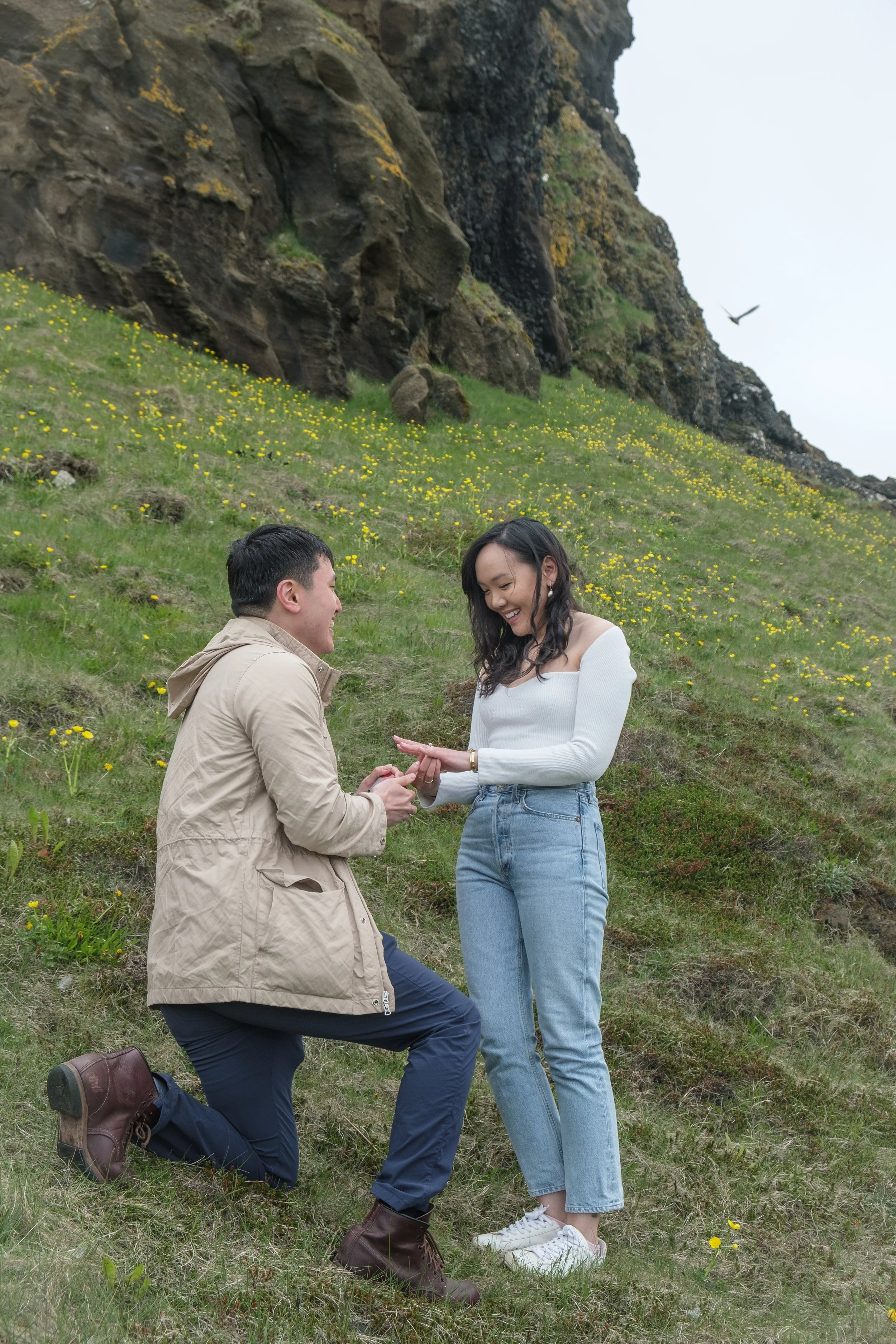 A couple is outside on a grassy hillside covered with small yellow flowers, with a rocky cliff in the background. One person is kneeling and proposing, holding the hand of a woman who is smiling. The woman is wearing a white off-shoulder top and ligh