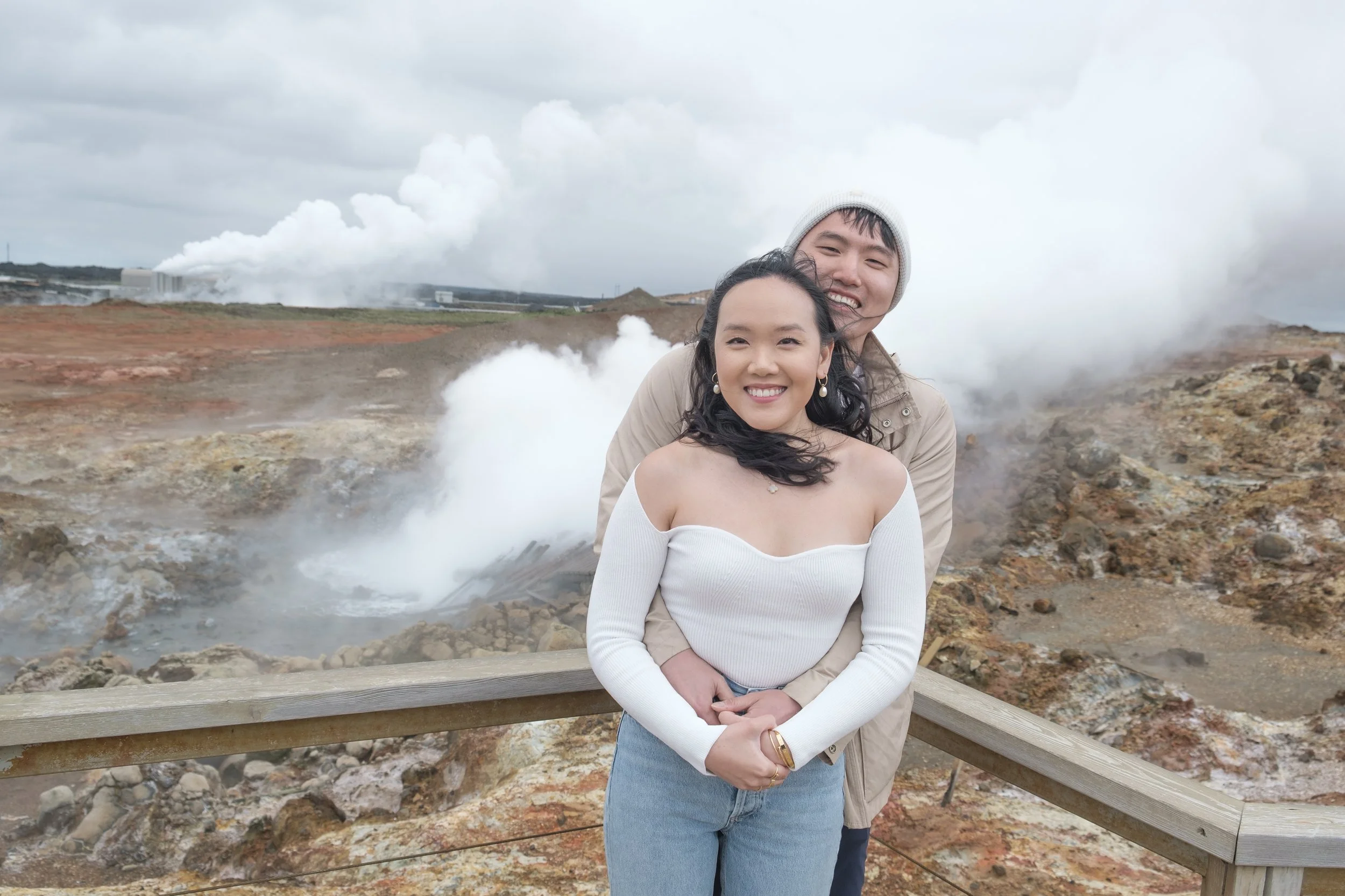 Two smiling people, a woman in a white off-the-shoulder top and a man wearing a white knit hat, standing near a wooden railing with geothermal activity and steam in the background.