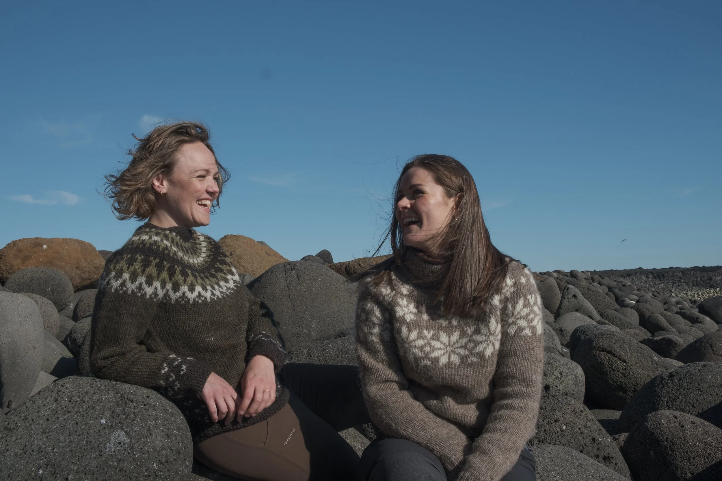 Two women sitting on rocks at the beach, smiling and talking, with a clear blue sky in the background.