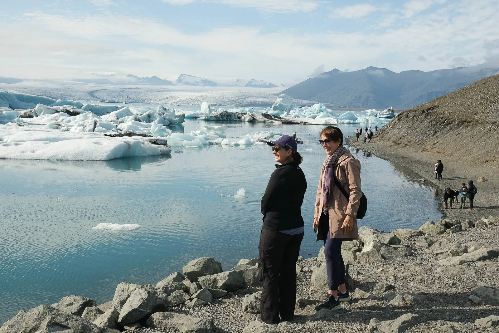 People standing on a rocky shoreline near a Glacier Lagoon & Ice Caves with floating ice chunks and distant mountains.