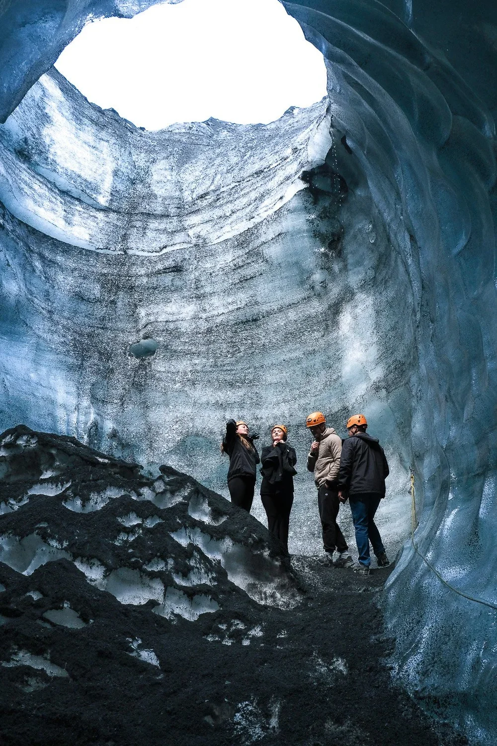 Four people wearing helmets stand inside a large ice cave with blue ice walls and black volcanic ash on the ground.