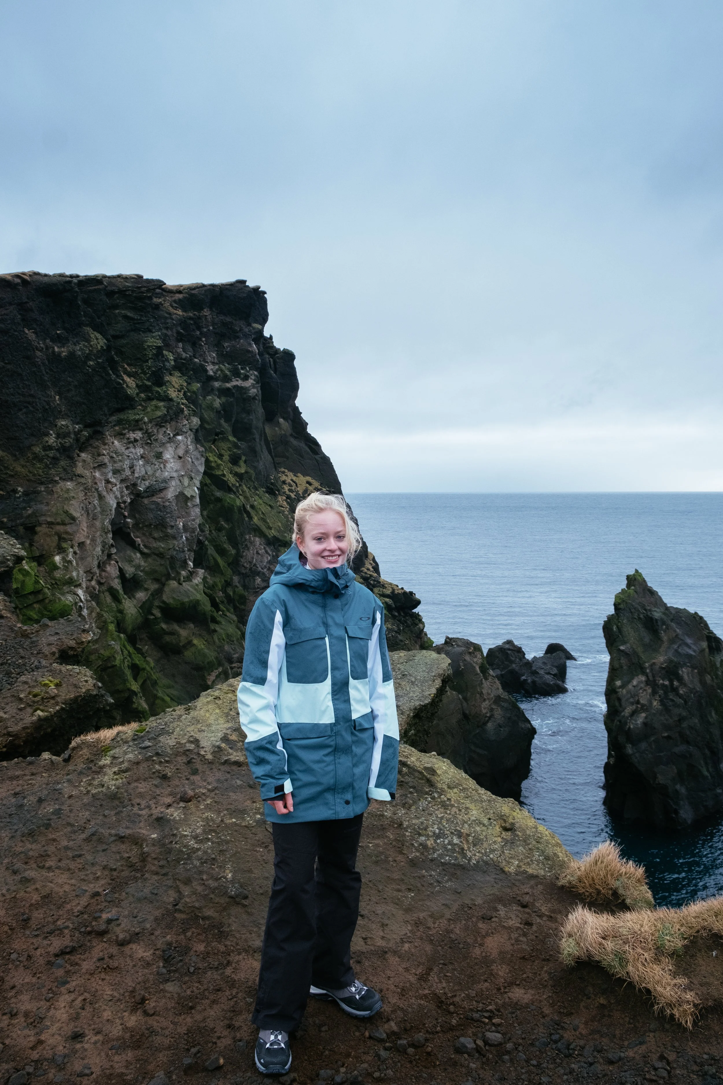 A smiling woman in a blue and gray outdoor jacket and black pants standing on rocky terrain near the ocean with green moss-covered cliffs and cloudy sky in the background.