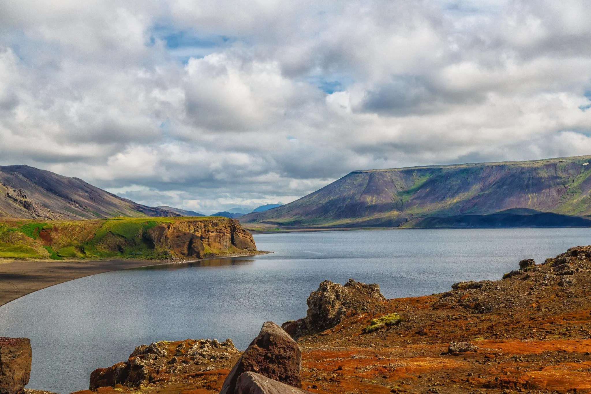 A scenic view of a Lake Kleifarvatn surrounded by green and rocky mountains under a sky filled with clouds