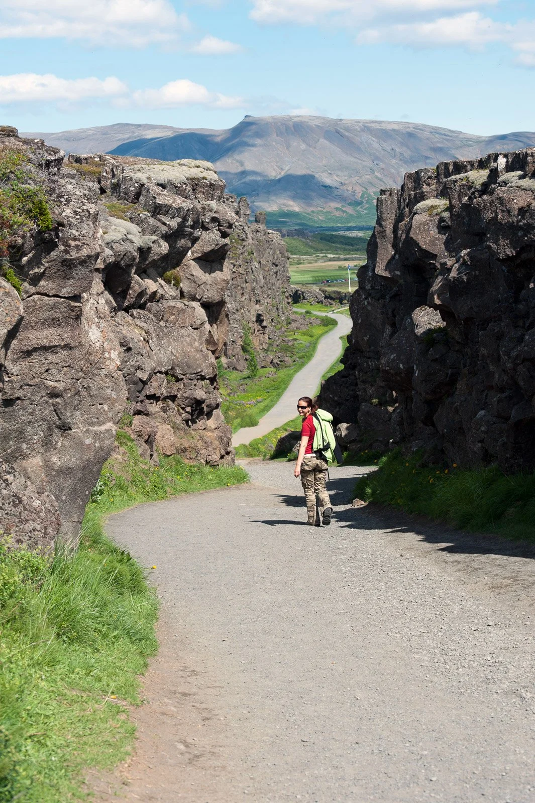 A woman hiking on a winding trail through a canyon with large rock formations on either side and a mountain in the distance under a partly cloudy sky.