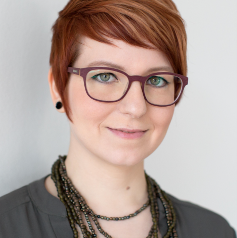 A woman with short red hair wearing glasses, a gray shirt, and layered necklaces, looking at the camera with a slight smile.