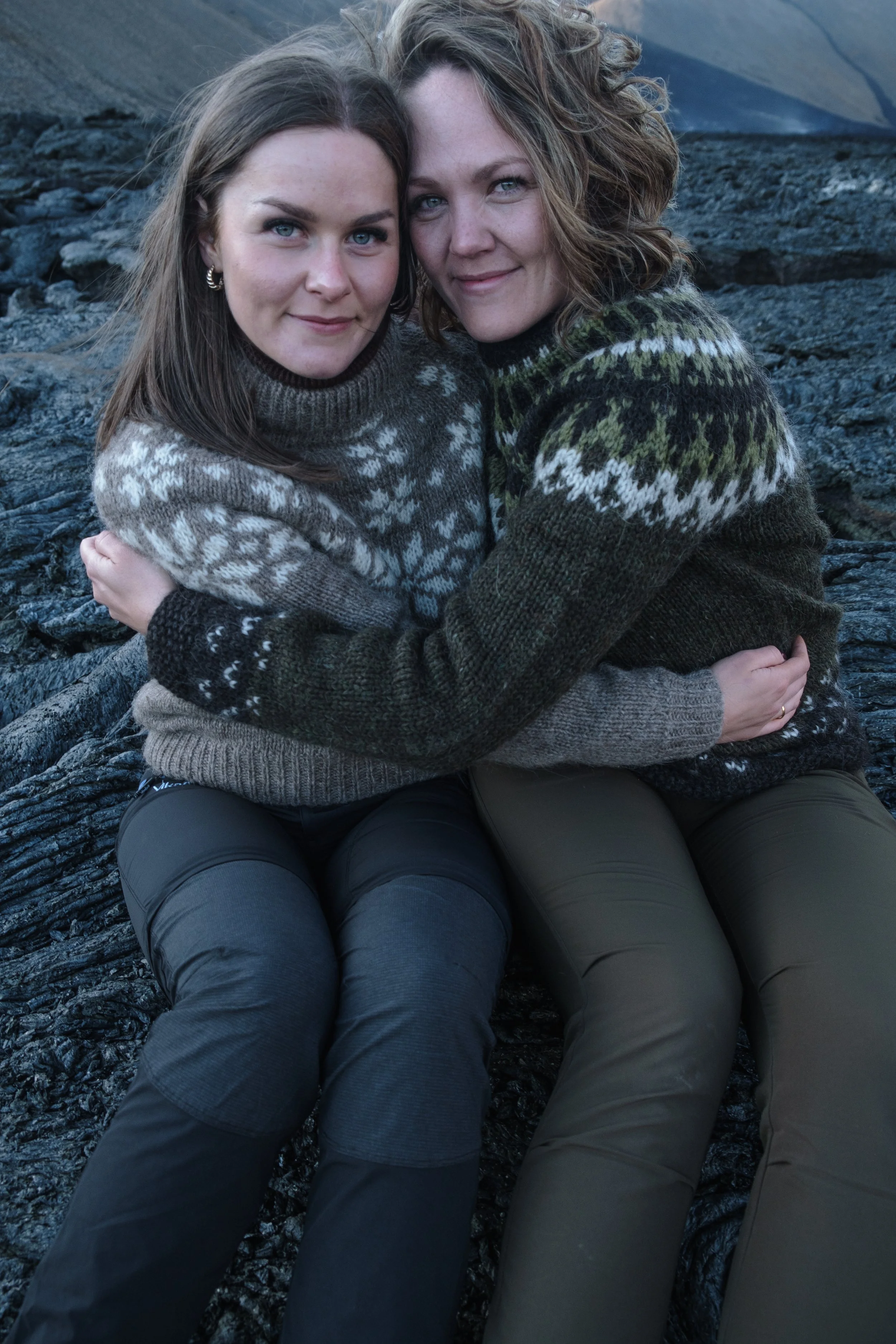 Two women sitting on rocks outdoors, wearing winter sweaters and smiling.