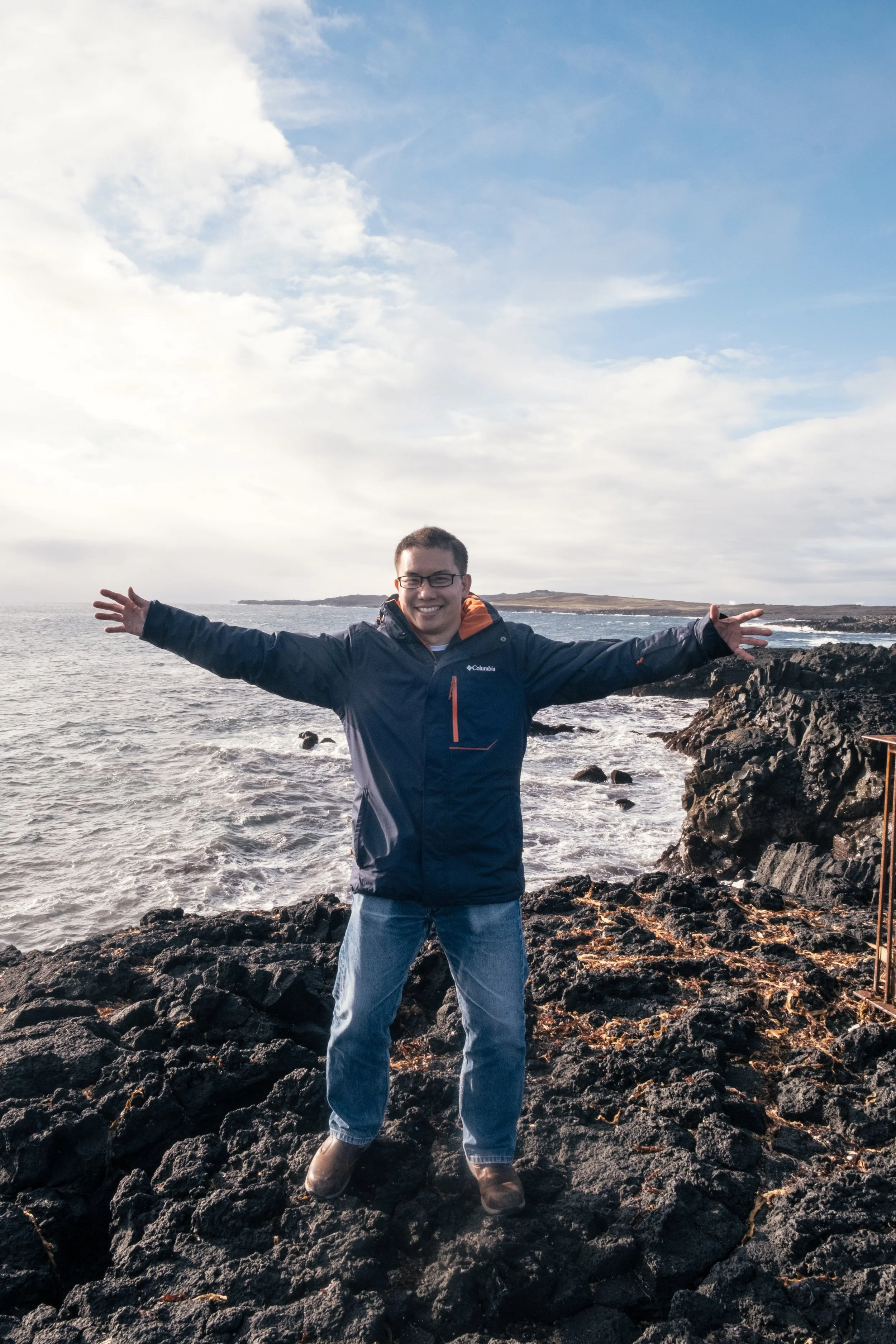 A man standing on rocky shore during daytime, arms outstretched, smiling, wearing a blue Columbia jacket, jeans, and boots, with ocean and coastline under partly cloudy sky in the background.