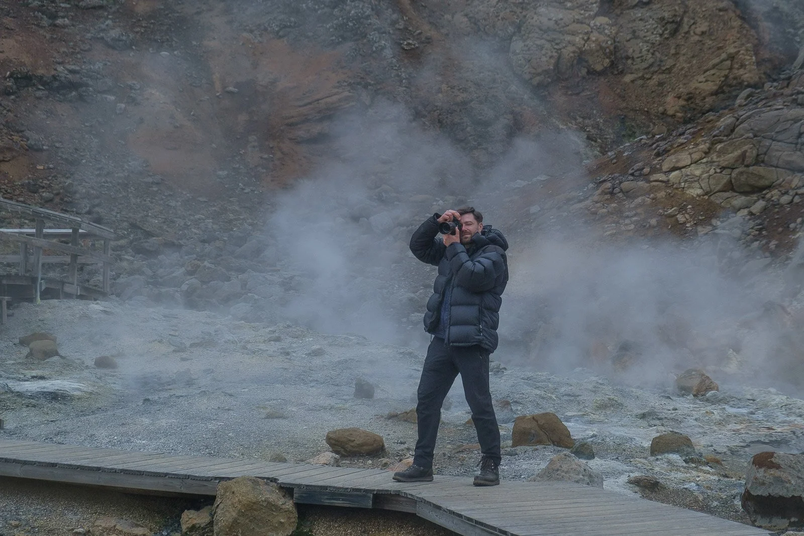 A man standing on a wooden boardwalk in a volcanic crater, holding a camera and taking a photo. The area is smoky and rocky with a rugged landscape in the background.