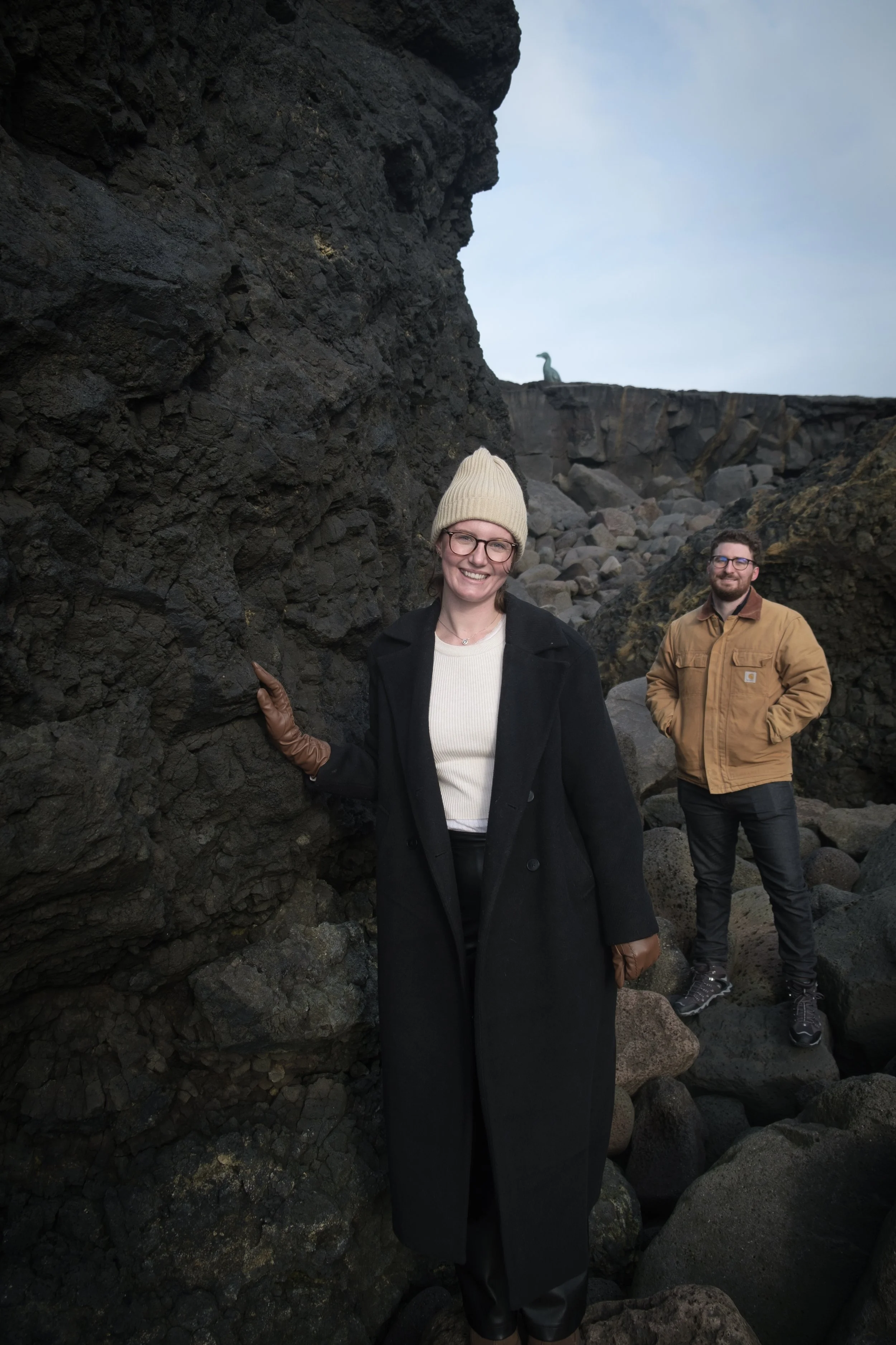 Two people standing near large dark rocks with a cliff and a dinosaur sculpture in the distance, on a rocky island or shoreline.