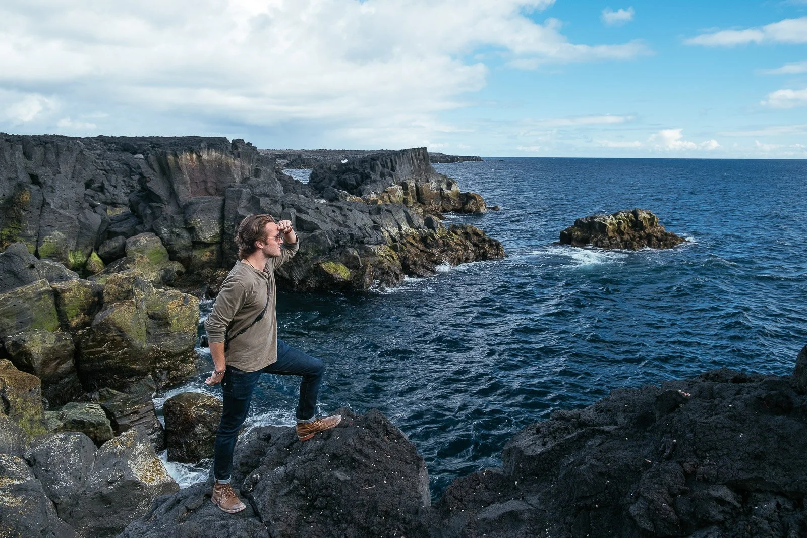 A man standing on black volcanic rocks by the ocean, looking out at the water with one hand on his forehead and the other behind his back, wearing sunglasses, a brownish-gray jacket, dark jeans, and brown boots.