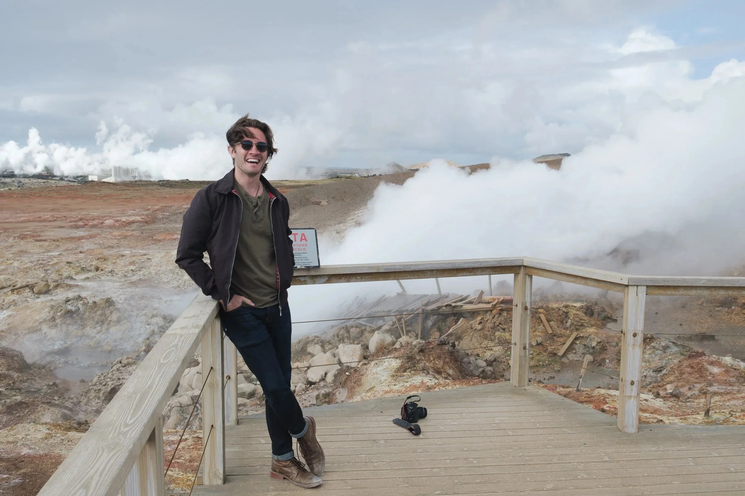 A young man with dark wavy hair, wearing sunglasses, a black jacket, and boots, smiling and standing on a wooden observation platform near geothermal steam and bubbling ground, with a cloudy sky in the background.