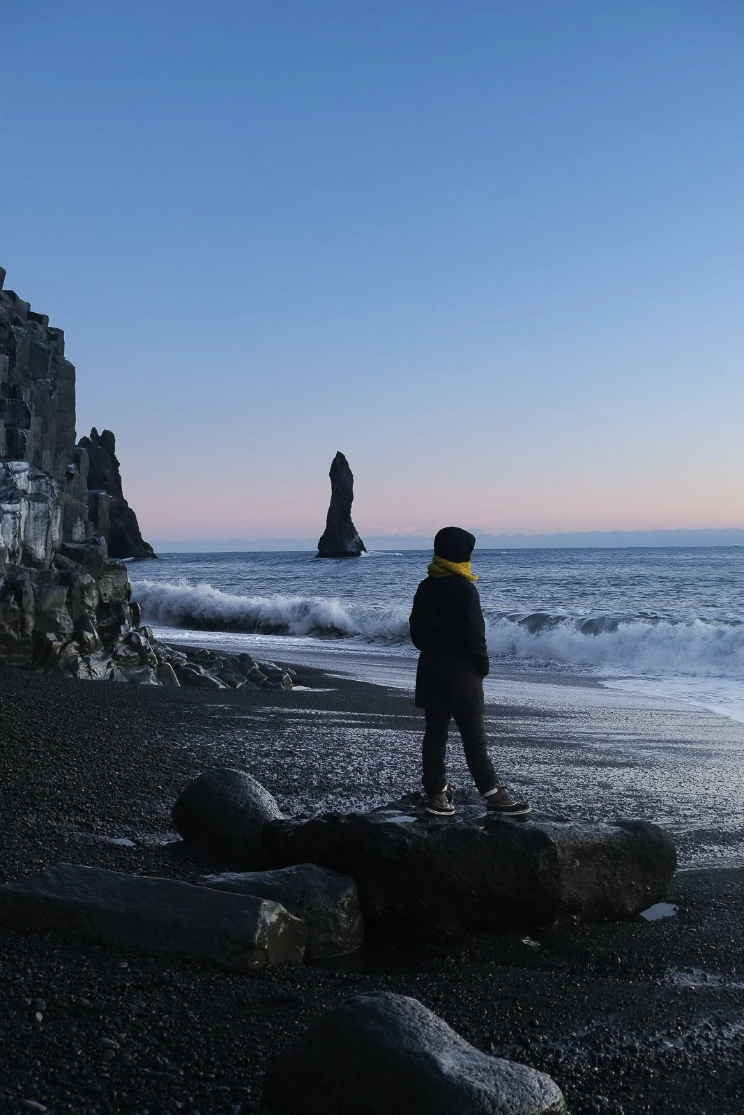 A person standing on a large rock on a beach, looking at the ocean and a tall sea stack formation during sunset or dawn.