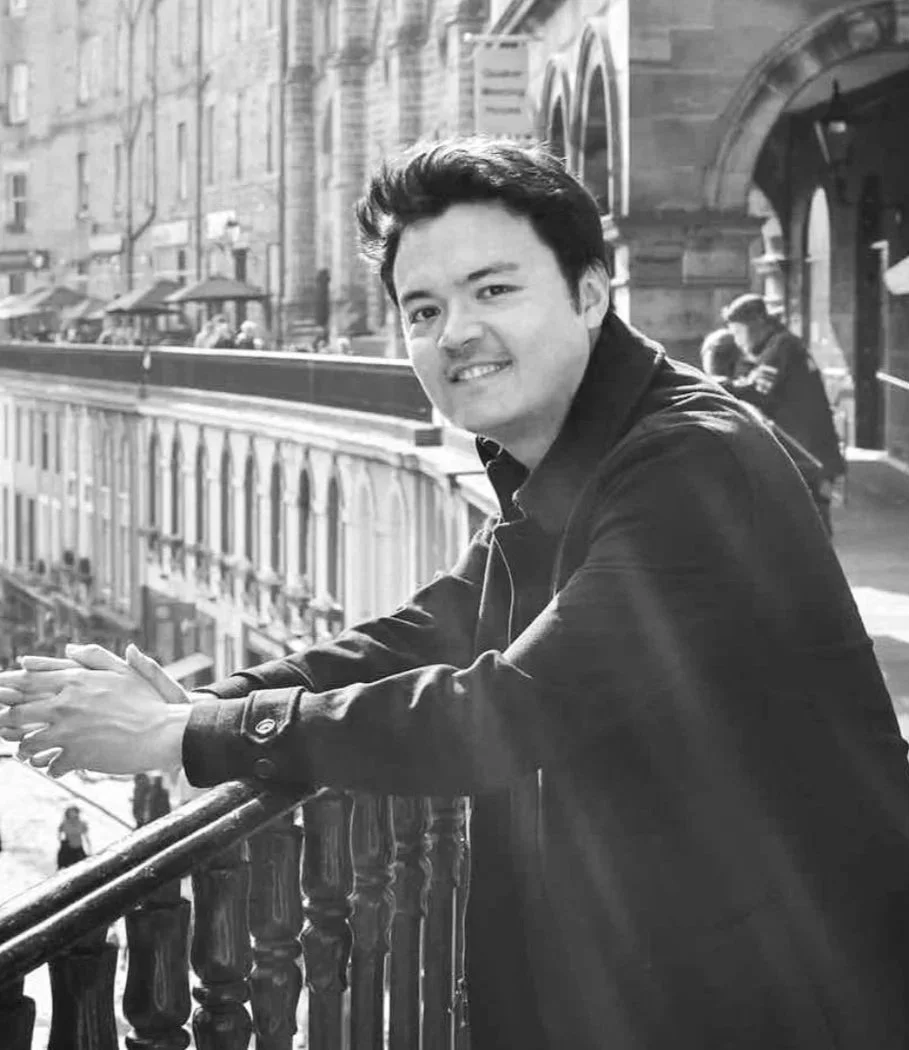 A young man leaning on a railing outdoors in an urban setting, with buildings and other people visible in the background.