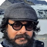 Close-up of a man with curly dark hair and beard, wearing dark sunglasses, a gray flat cap, and a navy blue jacket outdoors with snow-covered rocks in the background.