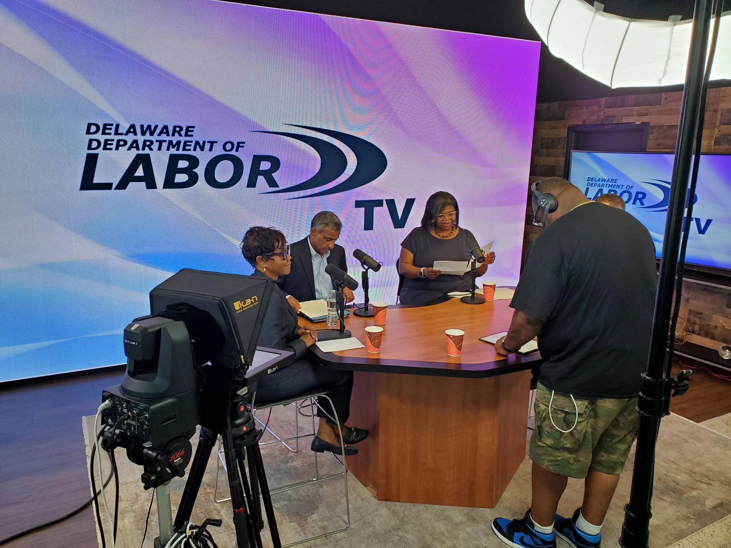 A behind-the-scenes shot of a live recording session for Delaware Department of Labor TV, featuring four people in a studio with a large digital screen displaying the department's logo. Two women and one man are seated at a curved wooden table with m