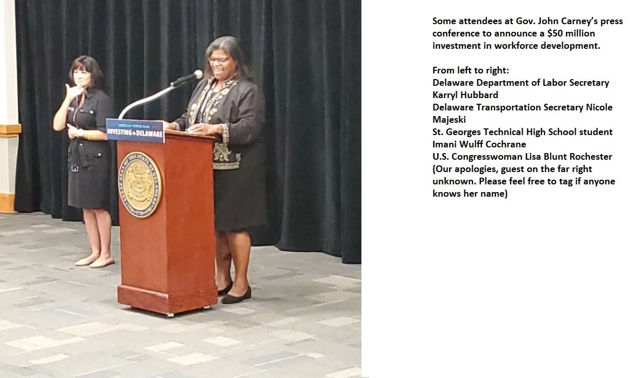 Several women standing on a stage, one at a podium speaking, others waiting to speak or observe, with black curtains behind them.