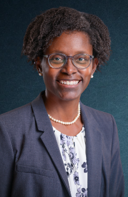 Professional woman with short curly hair, glasses, wearing a navy blazer, pearl necklace, and a floral blouse, posing against a dark teal background.