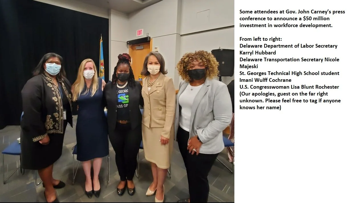 Group of five women standing together at a press conference, all wearing masks, with a black curtain and a flag in the background.