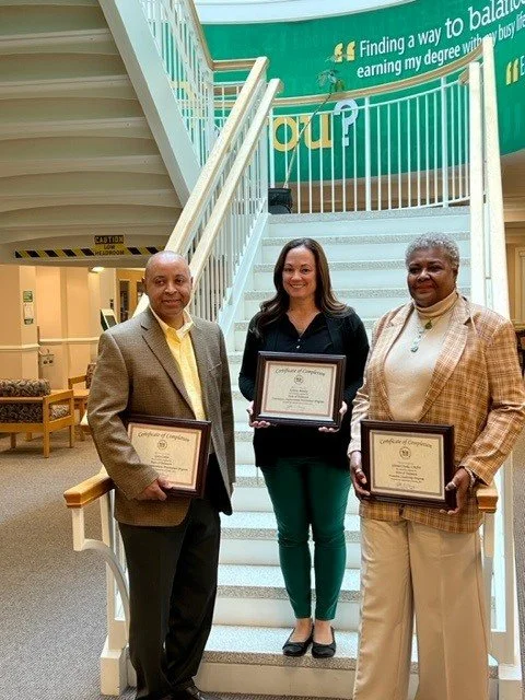 Three people standing on a staircase holding framed certificates, with a green banner in the background.