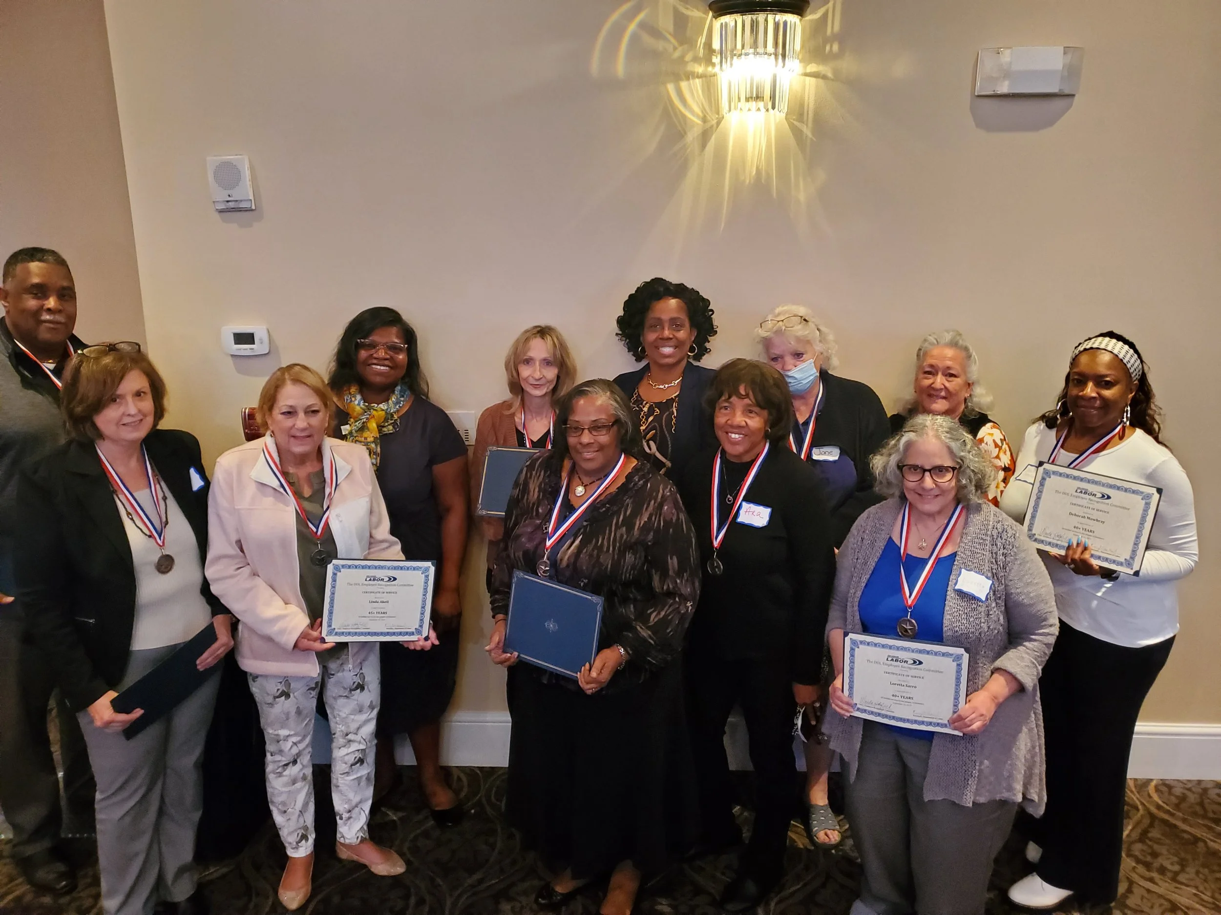 Group of diverse women posing together, some holding certificates and wearing medals, at an indoor awards or recognition event.